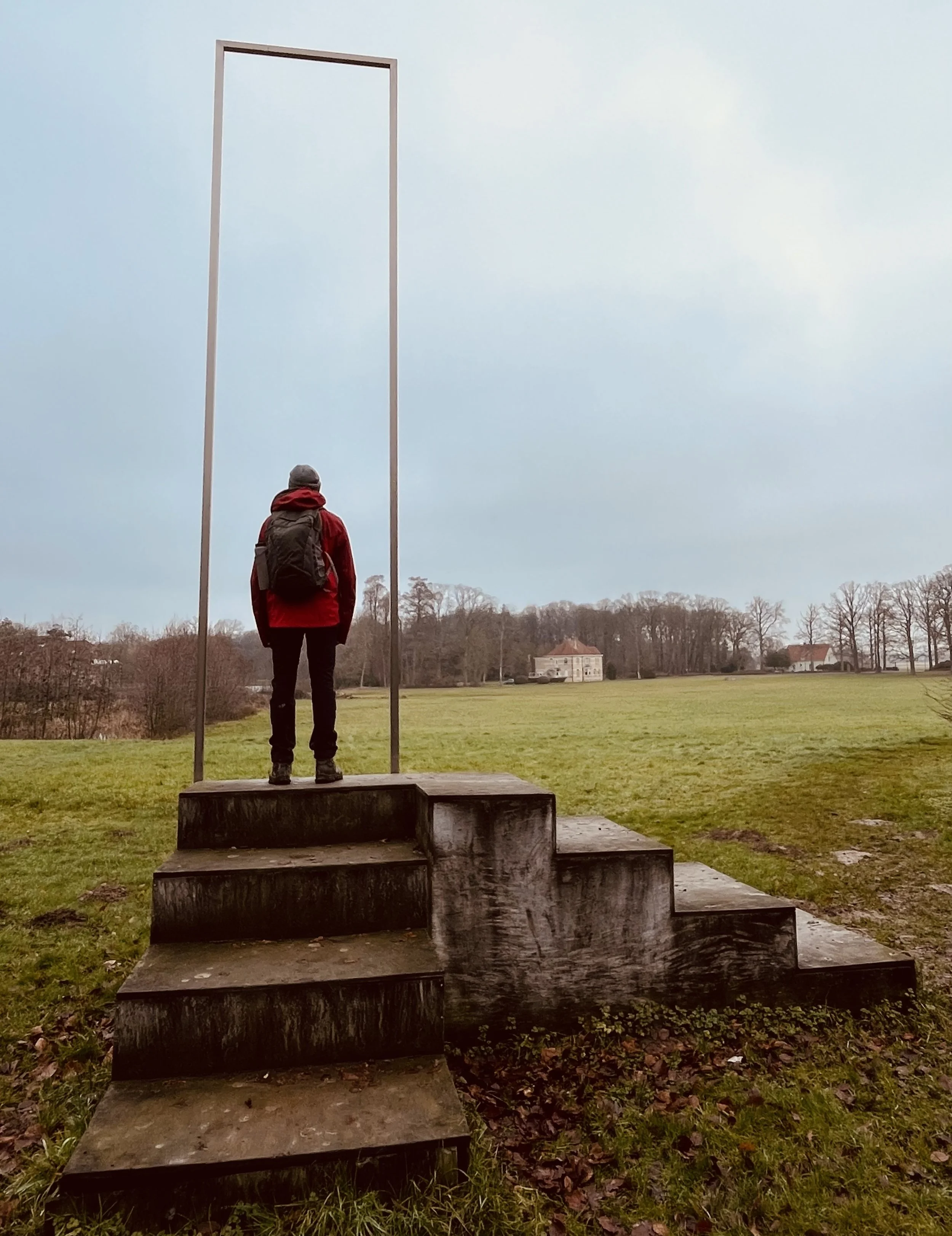 Persoon in een rood jack en rugzak kijkt uit over een uitgestrekt landschap met een historisch gebouw op de achtergrond en een grote, houten structuur met een open frame er boven.