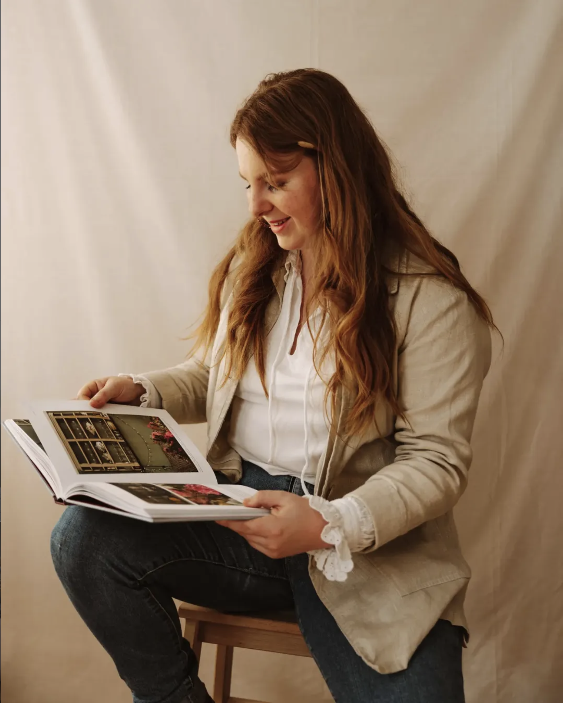 Une femme aux cheveux roux, assise sur une chaise, lit un livre ou un magazine dans un intérieur au mur clair.