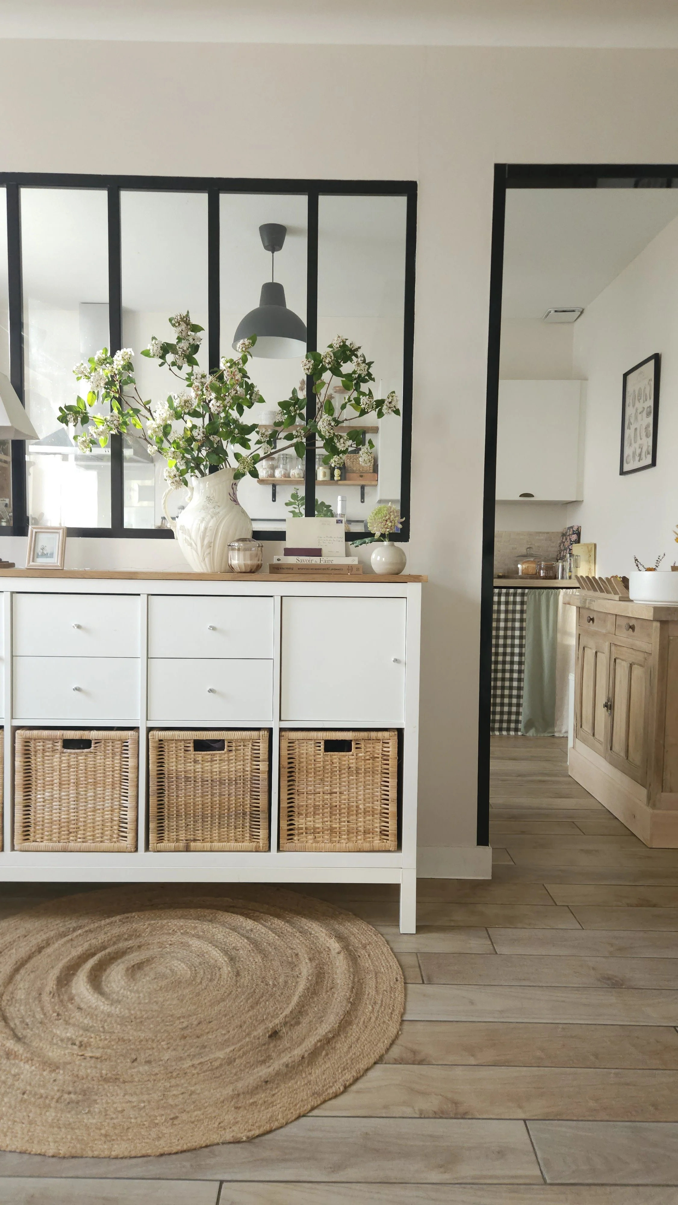 Intérieur de cuisine designé par Elsa Meslet, architecte d'intérieur à Cherbourg, avec un buffet blanc, fleurs dans une grande vase, et un tapis rond en jute sur le sol en bois.