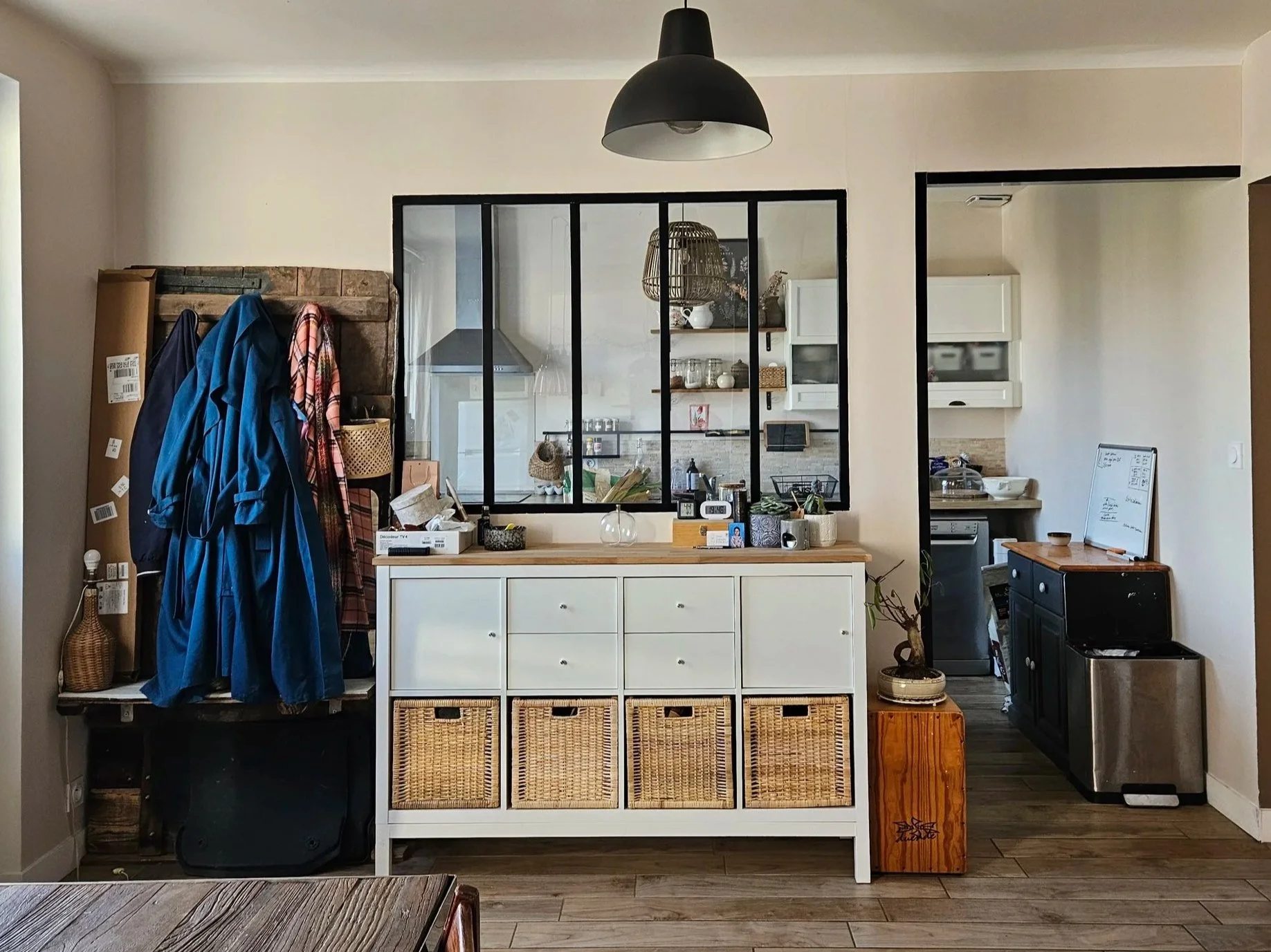 Une salle de séjour conçue par Elsa Meslet, architecte d'intérieur à Cherbourg, avec un meuble blanc contenant des paniers en osier, une verrière atelier et plusieurs objets décoratifs et utilitaires sur le meuble et le comptoir