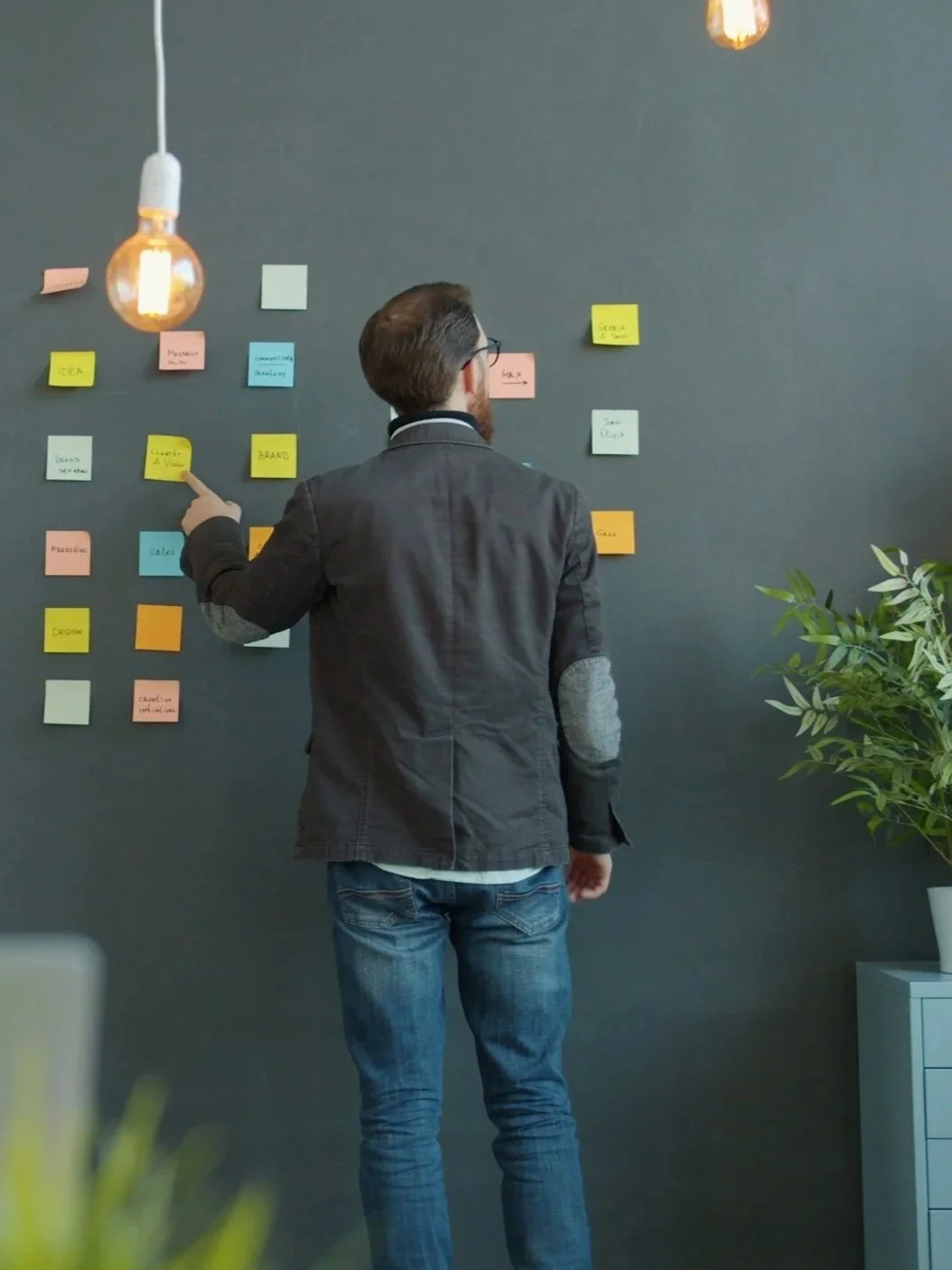 A man in a black blazer and jeans standing in front of a black wall with colorful sticky notes, pointing at one of the notes. Hanging light bulbs and a potted plant are also visible.