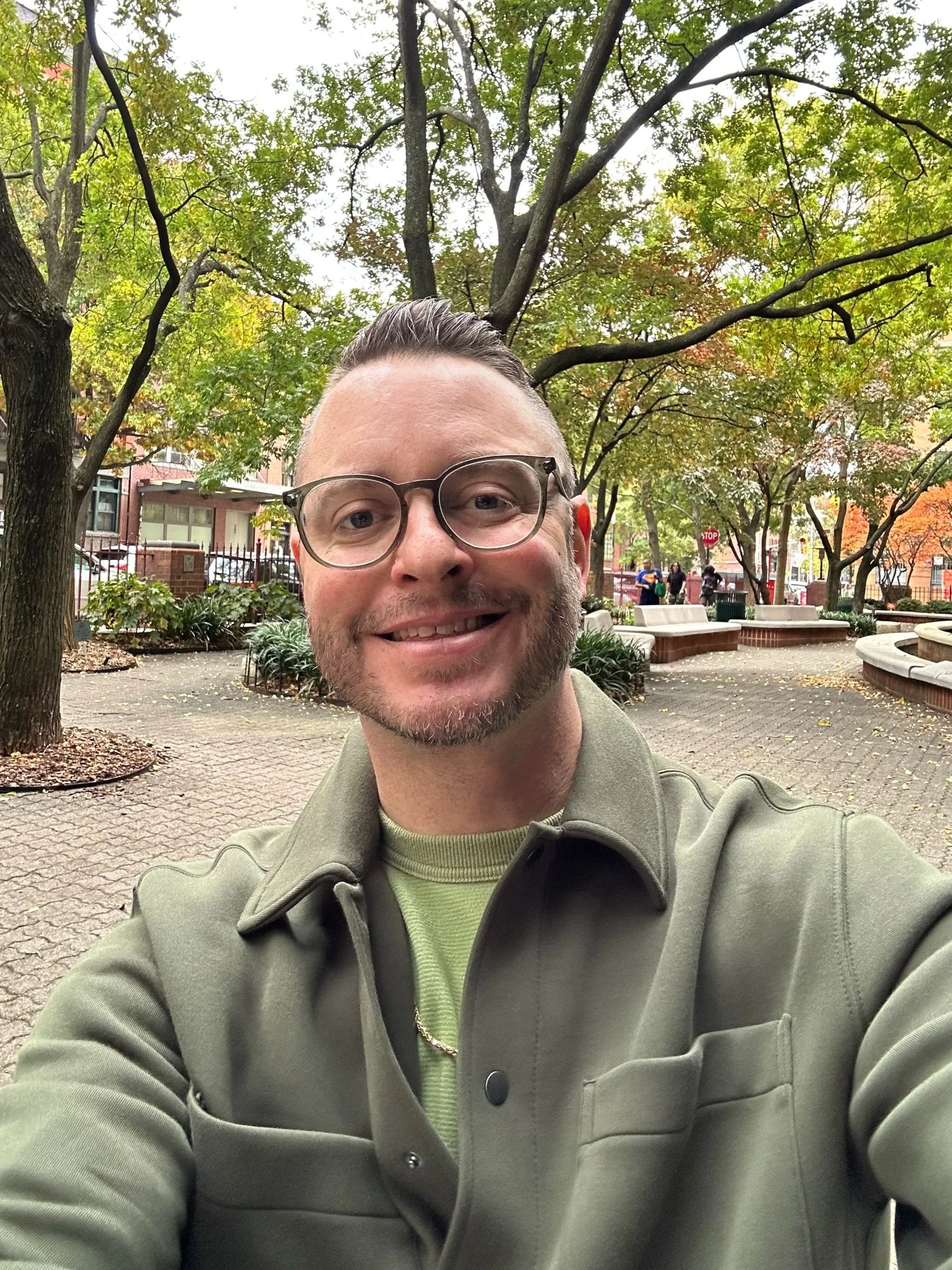 A man smiling and taking a selfie in a park with green trees and benches in the background.