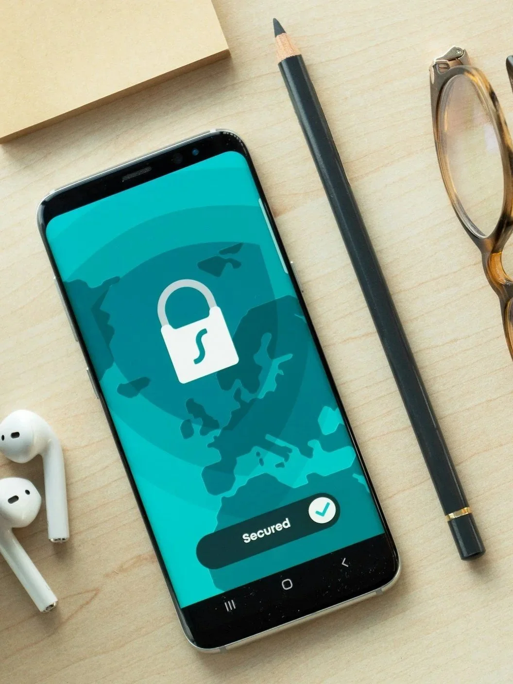 Smartphone on a desk displaying a security app with a lock icon and the word "Secured." Surrounding items include wireless earbuds, a black pencil, a pair of glasses, and a notebook.