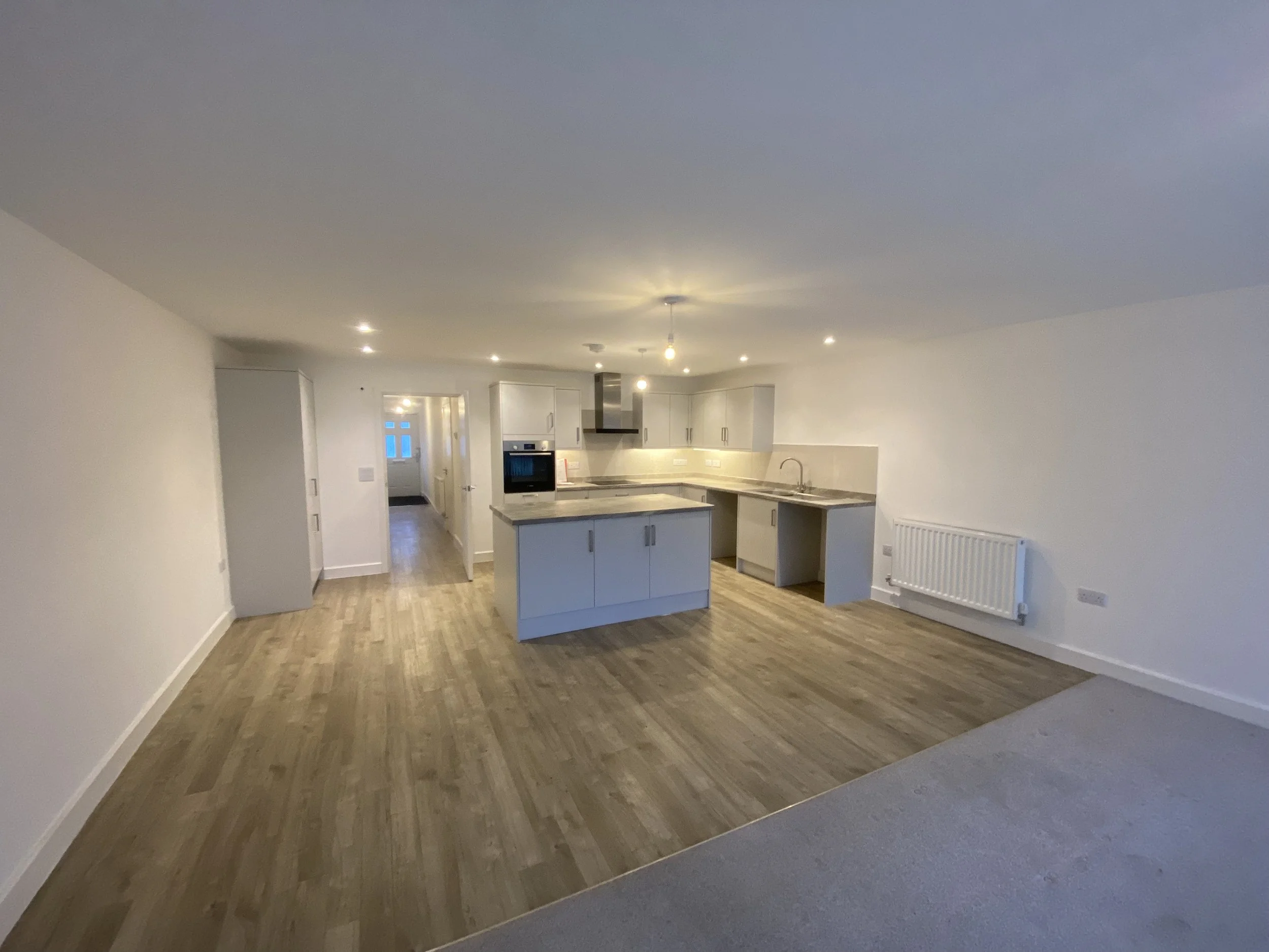 Empty open-plan kitchen and living space with white walls, wood flooring, and kitchen cabinets, oven, and sink on the right.