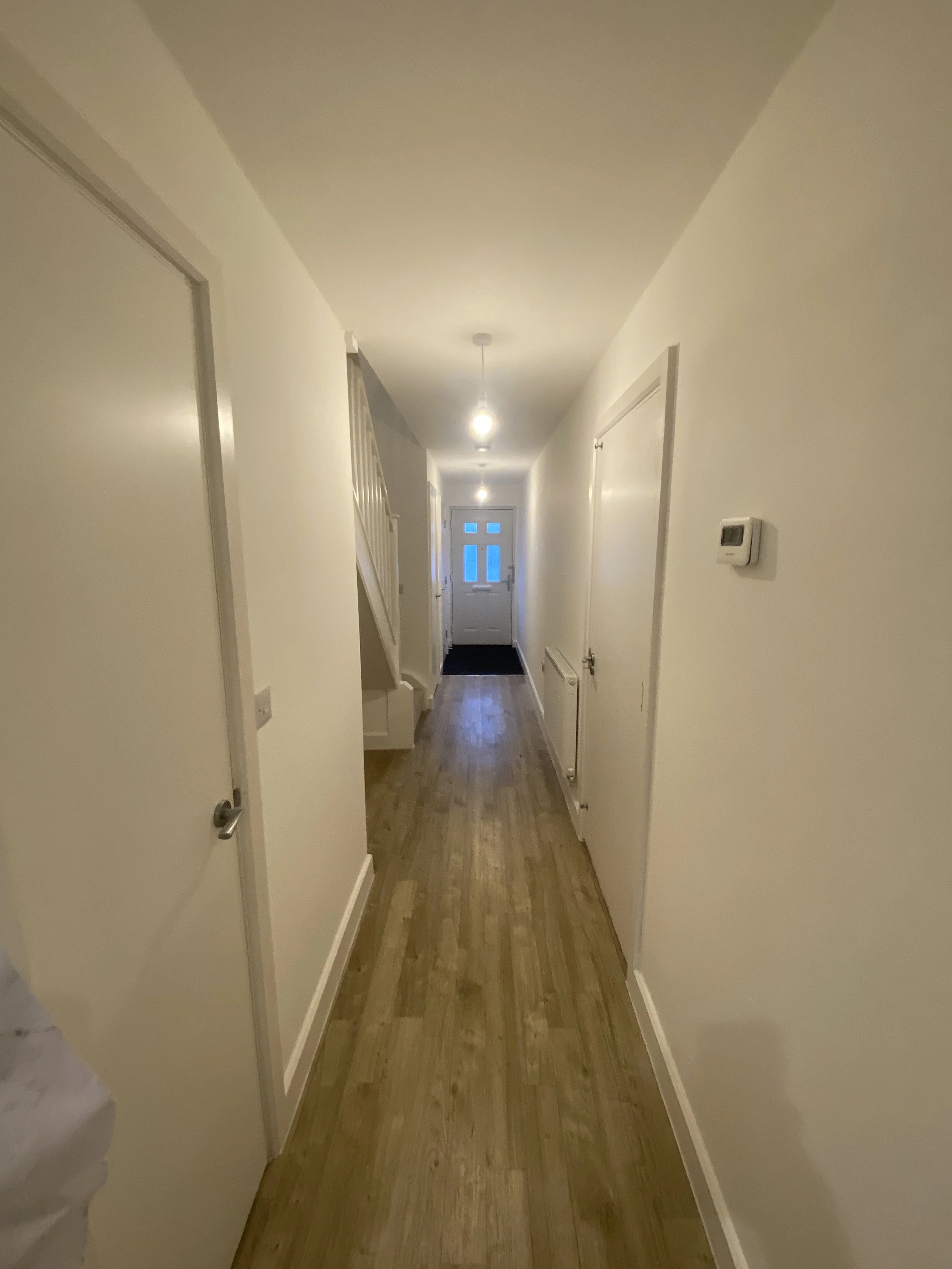 Empty hallway with white walls, wooden flooring, and ceiling lights leading to a door with a window at the end.