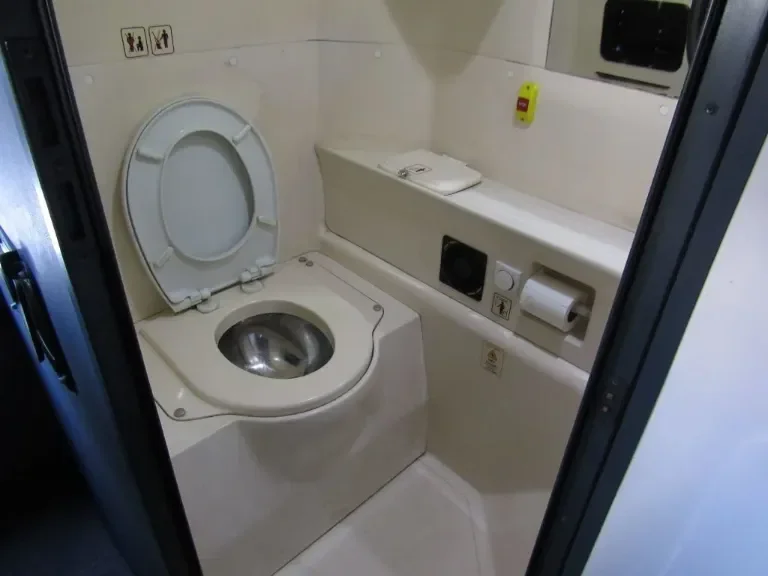 Small private bathroom with a toilet featuring a stainless steel seat and lid, and a wall-mounted shelf with a folded towel. The walls are light-colored, and there are various controls and fixtures nearby, including a paper towel dispenser.