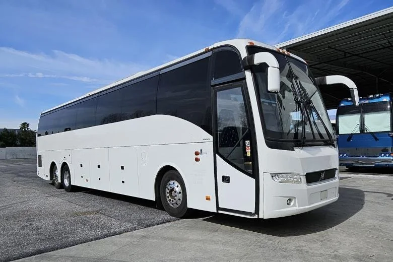 A large Volvo bus parked on a lot, with another bus visible in the background under a covered area.