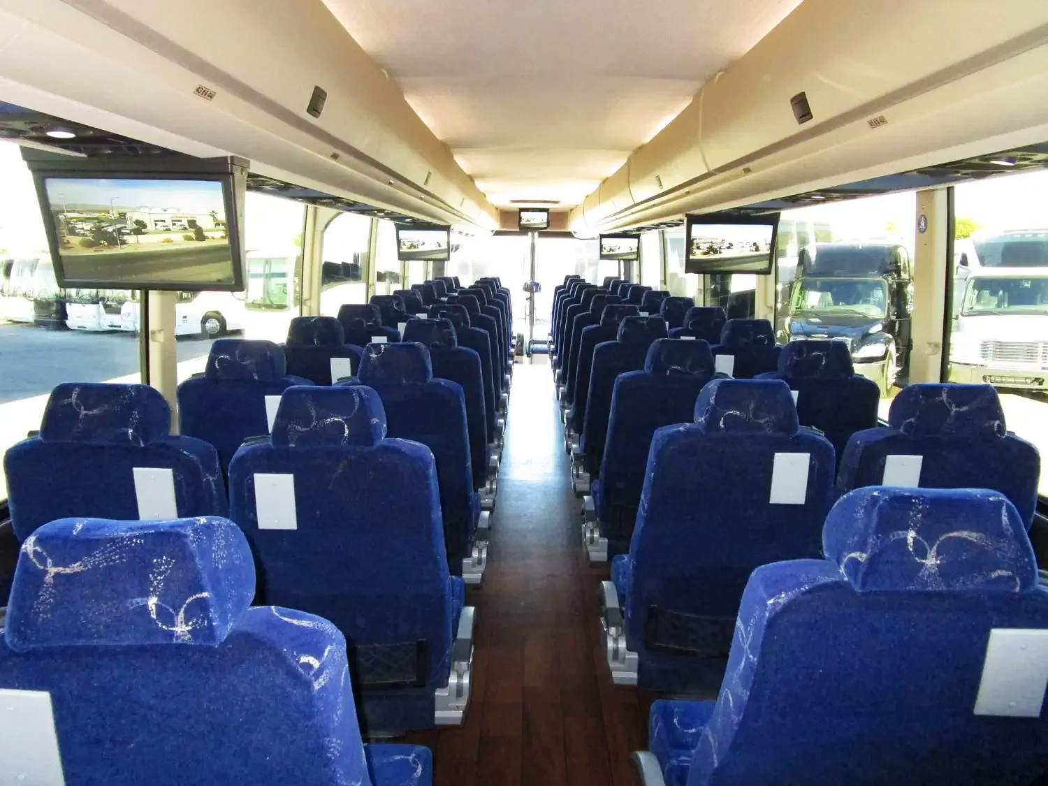 Interior of a bus with rows of blue seats facing forward and multiple screens mounted on the ceiling, with windows showing parked vehicles outside.