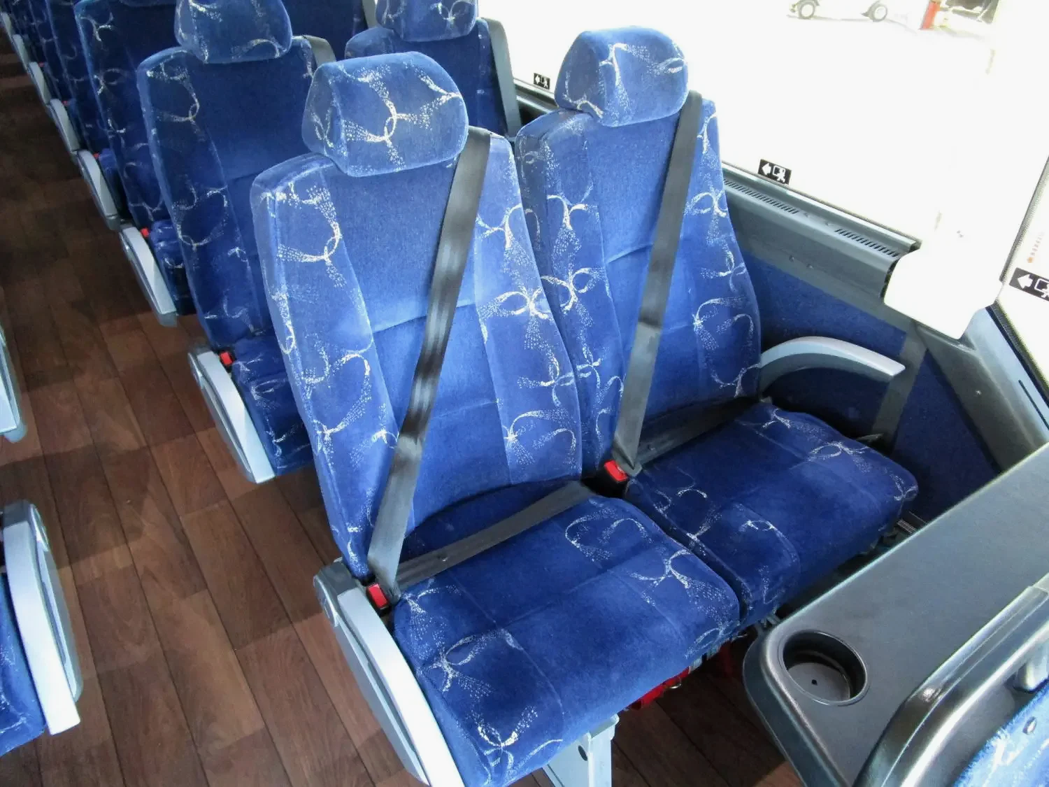 Several blue fabric bus seats with white swirl pattern, retractable seat belts, and a window on the side, on a wooden floor.