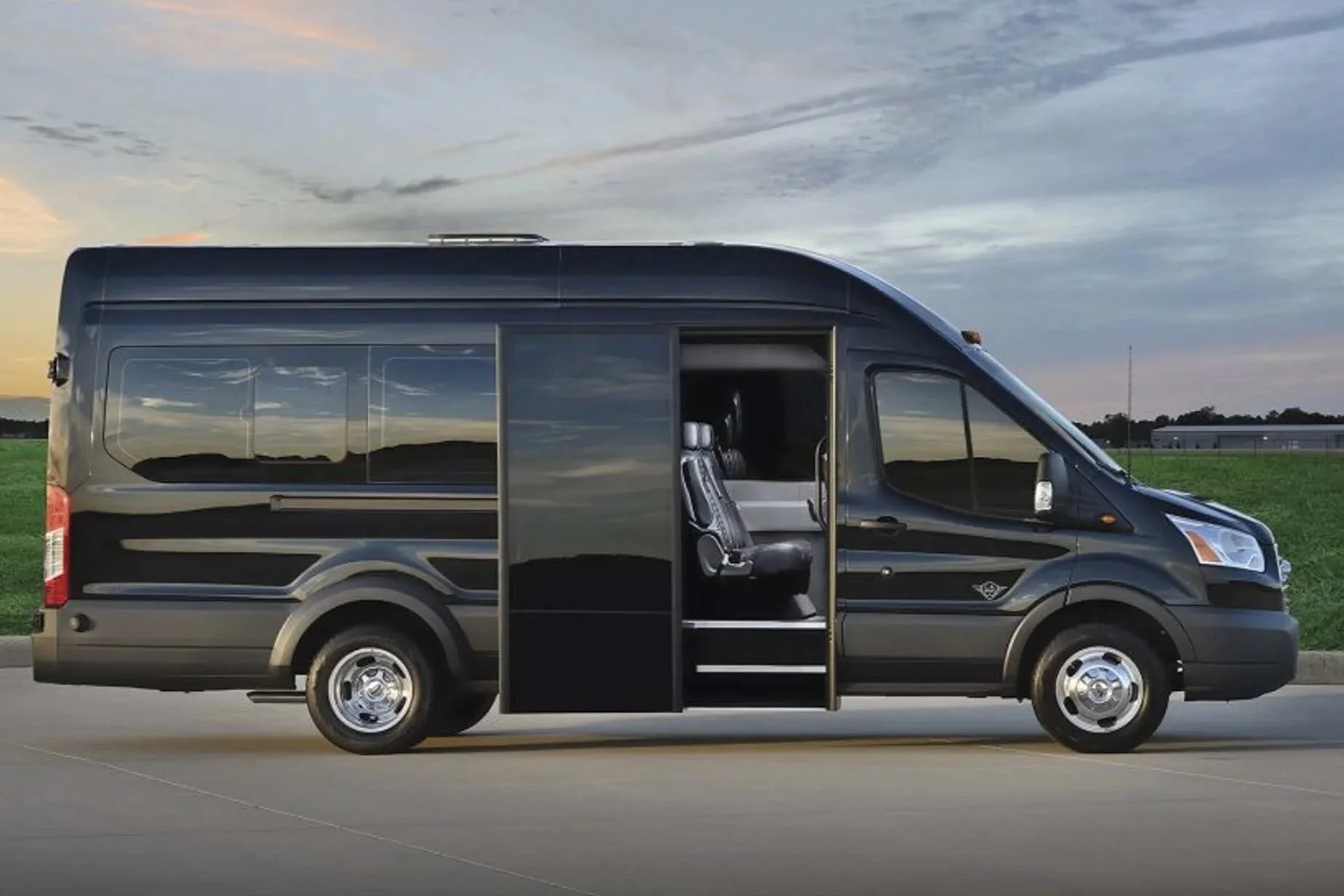 A black luxury Ford Transit van parked outdoors on a paved surface, with its sliding door open revealing seating inside and a scenic landscape with grass and a cloudy sky in the background.