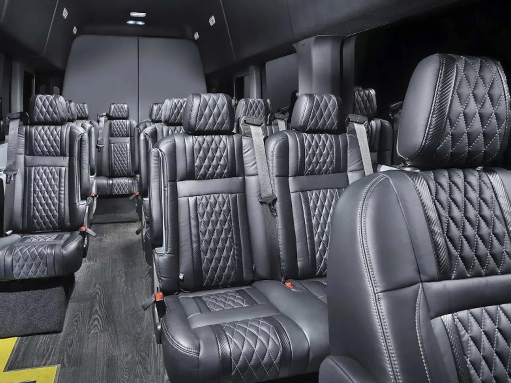 Interior of a luxury Ford Transit van with black quilted leather seats arranged in rows, with seat belts visible, and a dark ceiling with ceiling lights.