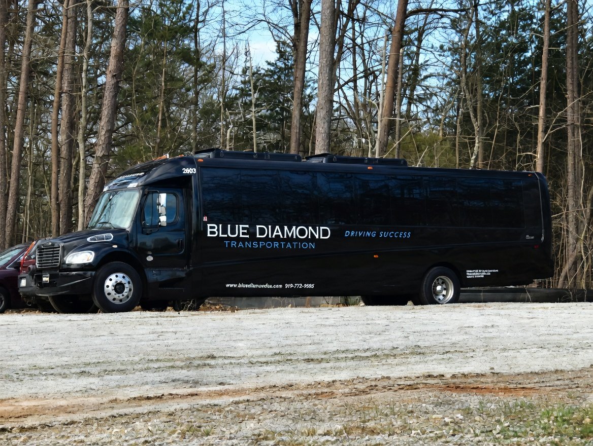 Black Frieghtliner Mini Bus with Blue Diamond company logo parked on a gravel road in front of a wooded area.