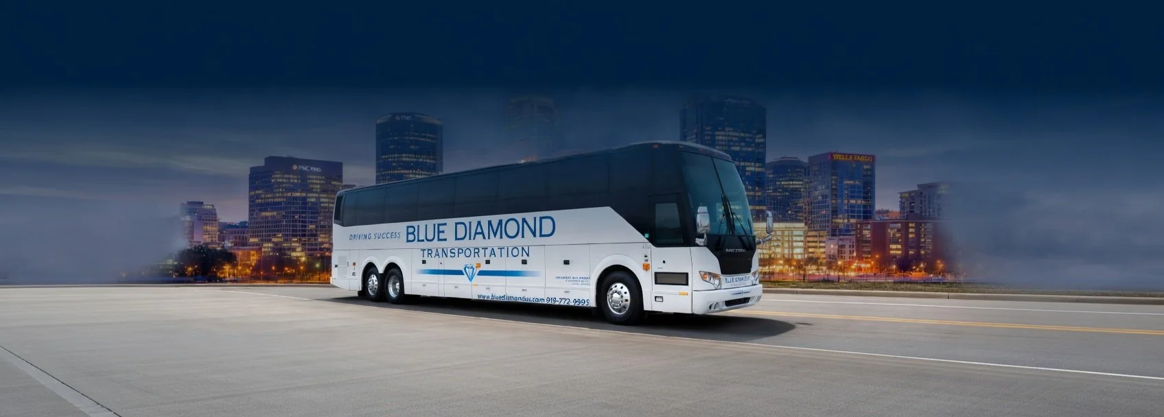 A white and black bus with blue lettering on the side reading 'Blue Diamond Transportation' driving on a city street at night, with a city skyline of illuminated buildings in the background.