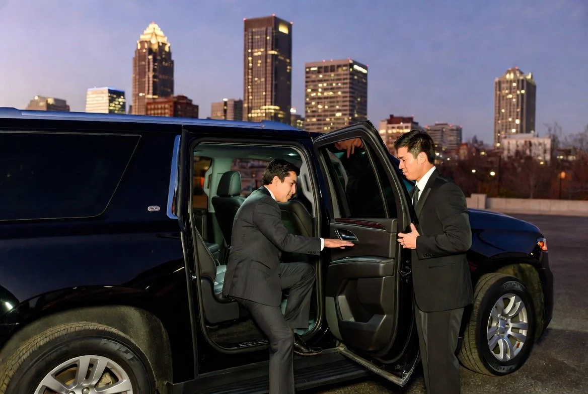 Two men in suits standing next to a black SUV with open doors, one is seated inside the car and the other is standing outside, in an urban cityscape at dusk.
