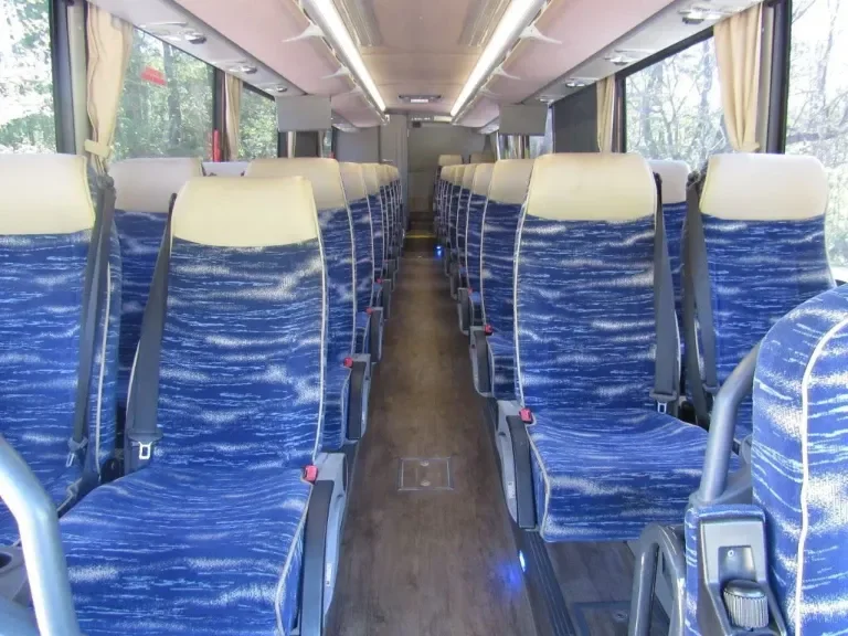 Interior of a bus with two columns of blue upholstered seats with cream headrests, aisle in the middle, and large windows with curtains.
