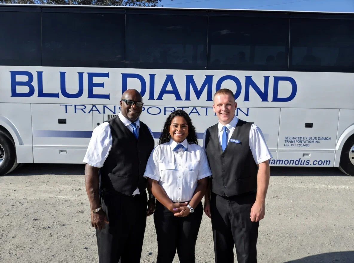Three smiling transportation employees standing in front of a Blue Diamond Transportation bus. The man on the left is wearing sunglasses, a white shirt, and a black vest. The woman in the middle is wearing a white blouse with a blue tie. The man on the right is wearing a white shirt with a black vest and a blue tie.