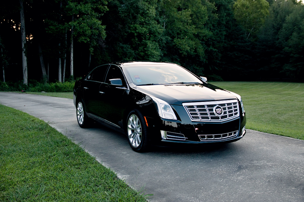 Black Cadillac XTS sedan parked on a narrow driveway next to grass and trees to pick up client.
