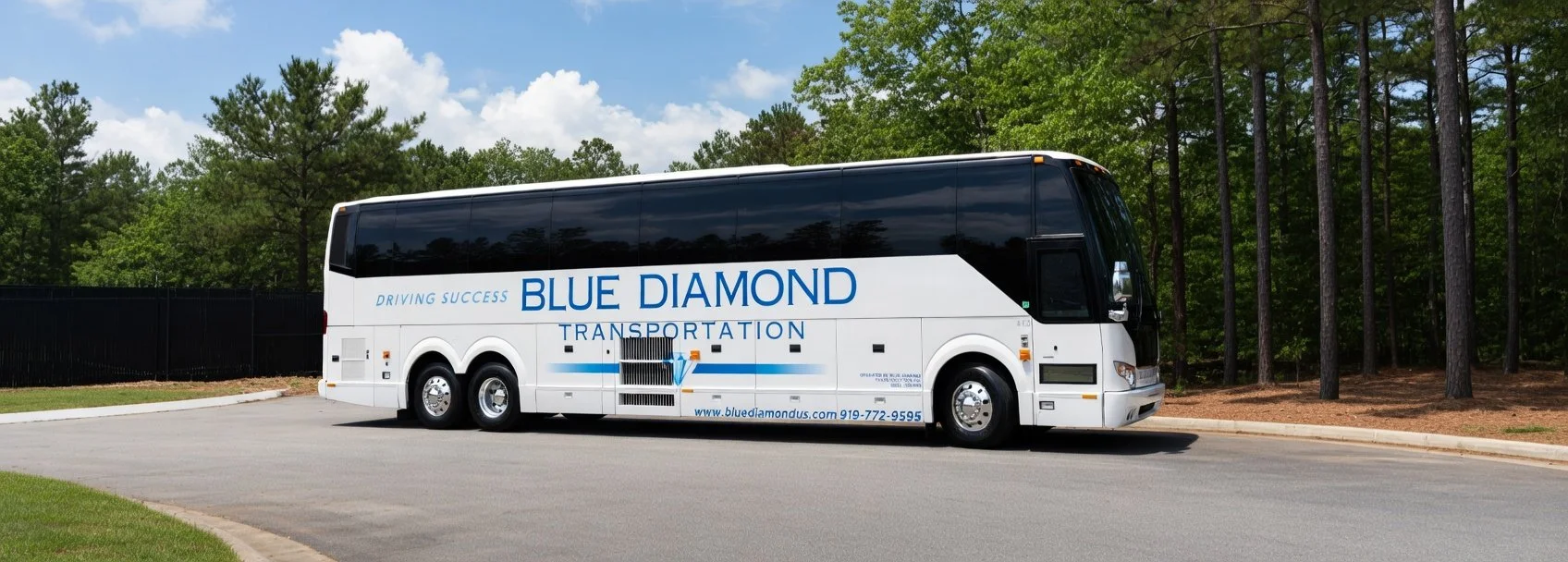 White Prevost Motor Coach with the Blue Diamond Transportation logo parked on a paved road next to a grassy area with trees in the background.