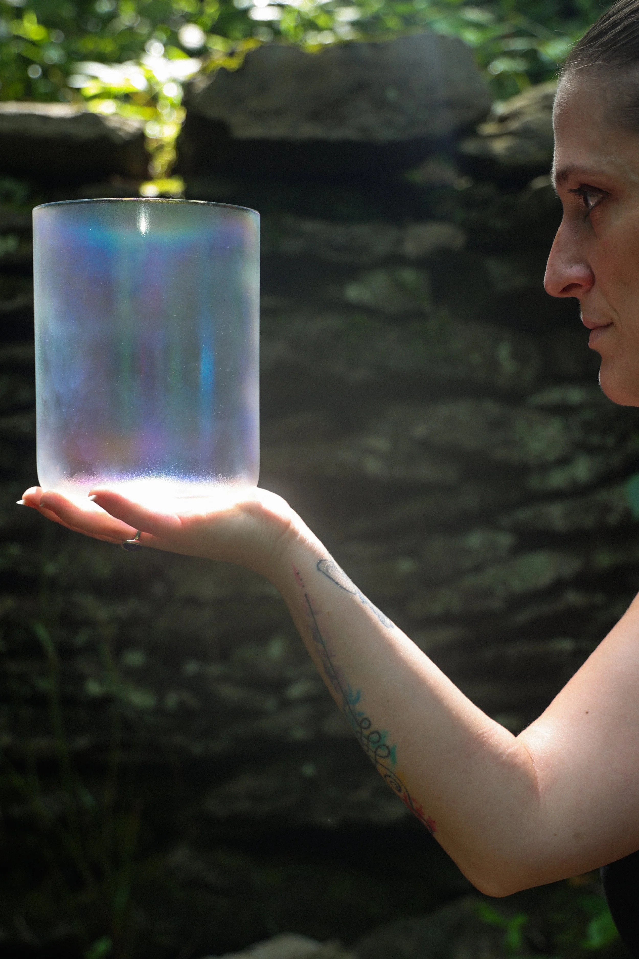 A person with tattoos on their arm holds a large iridescent crystal singing bowl in a natural outdoor setting with rocks and greenery.