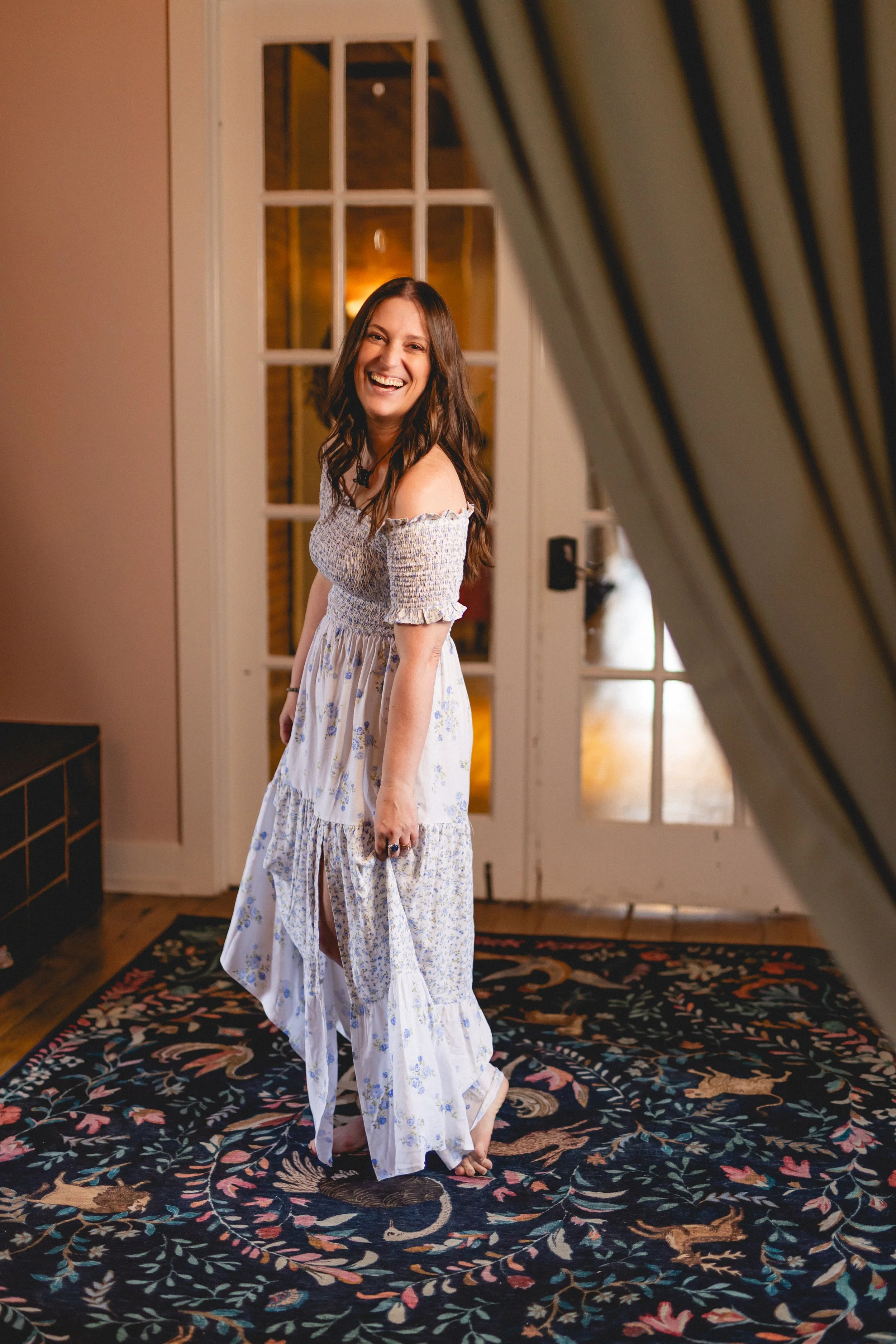 A woman with long brown hair, smiling and laughing, standing on a floral patterned rug inside a room with glass doors and yellow lighting.