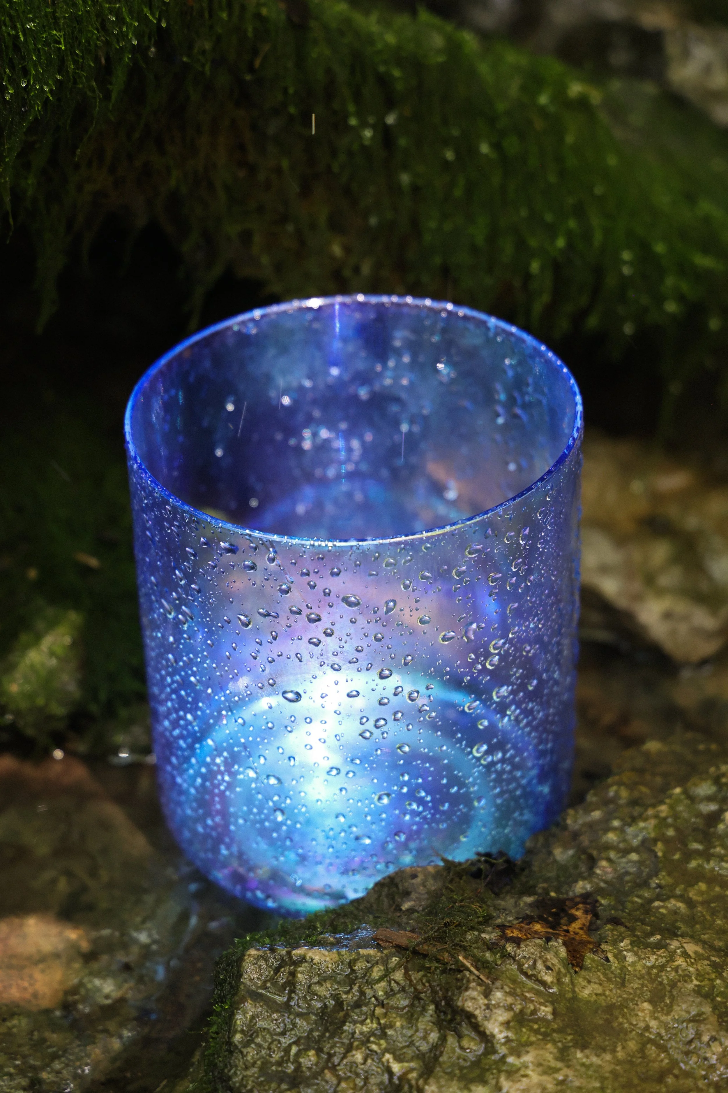 A blue crystal singing bowl with water droplets on the outside, placed on mossy rocks in a natural outdoor setting.