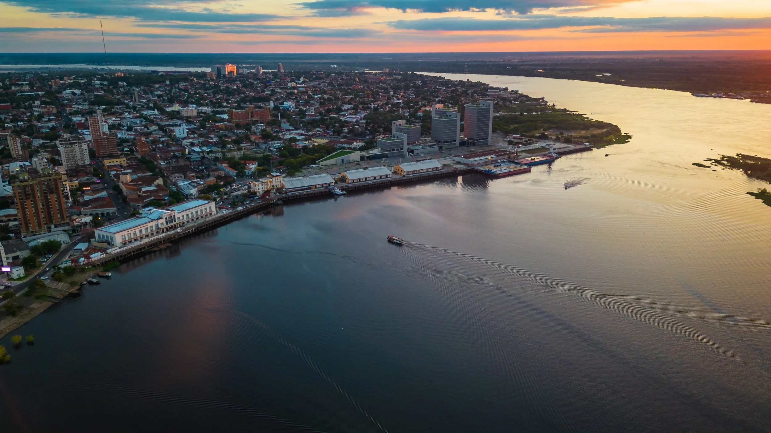 Aerial view of a city by a river at sunset, with boats on the water and a skyline including tall buildings and a mix of residential and commercial structures.
