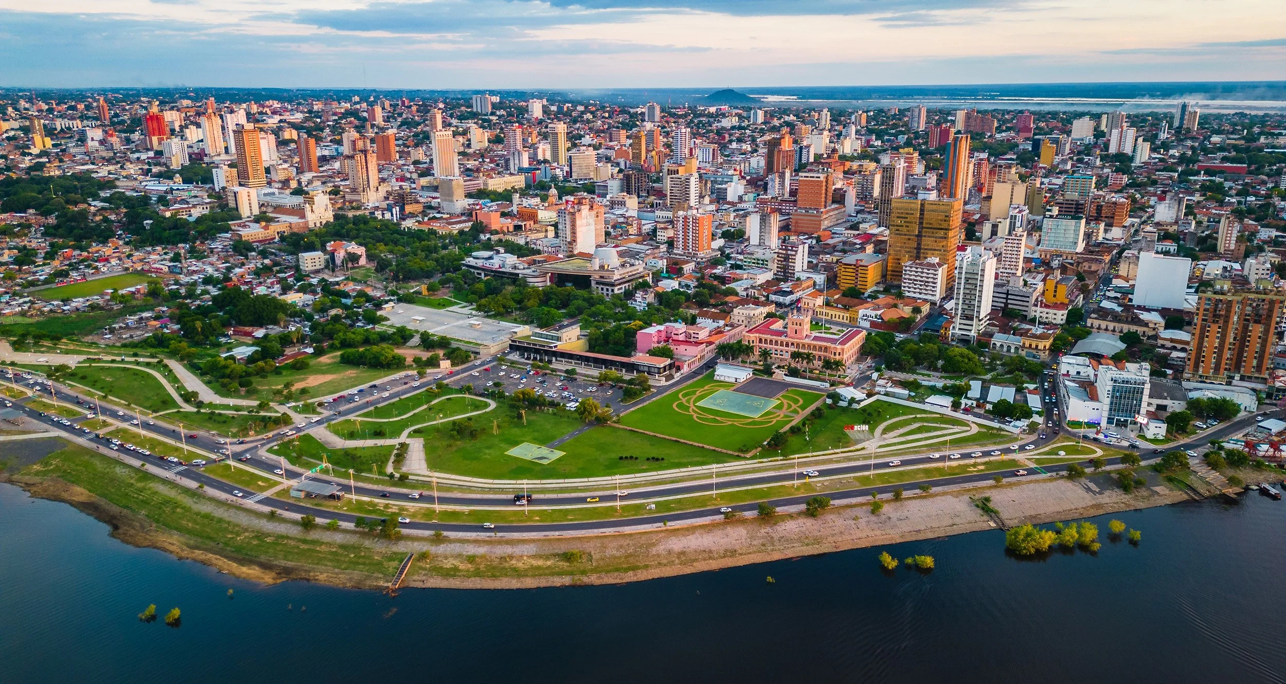 Aerial view of a city skyline with high-rise buildings, a river in the foreground, and a park with walking paths, tennis courts, and a pink historic building in the middle.