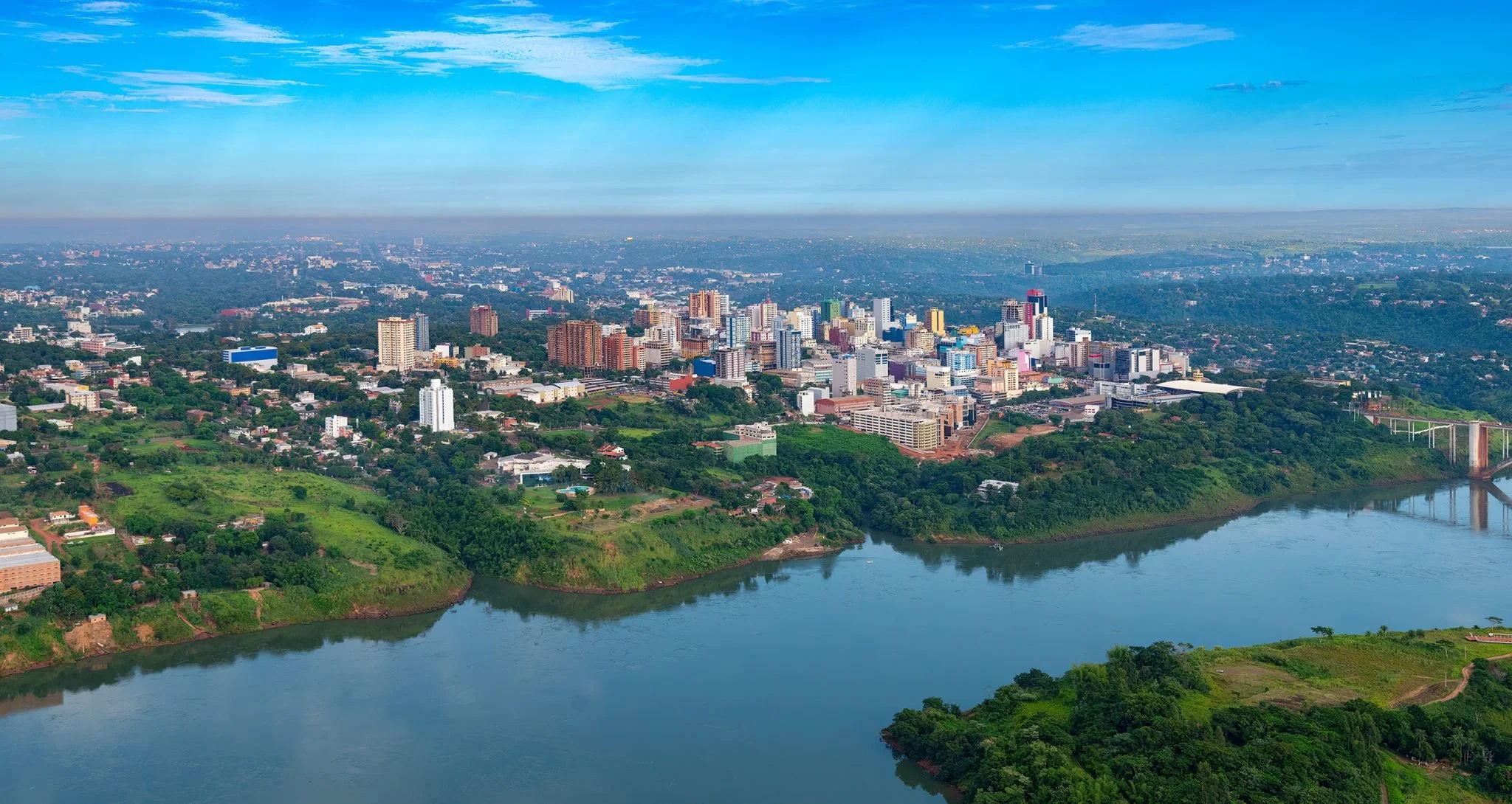 Aerial view of a city with numerous buildings, a river in the foreground, green hills, and a clear blue sky.