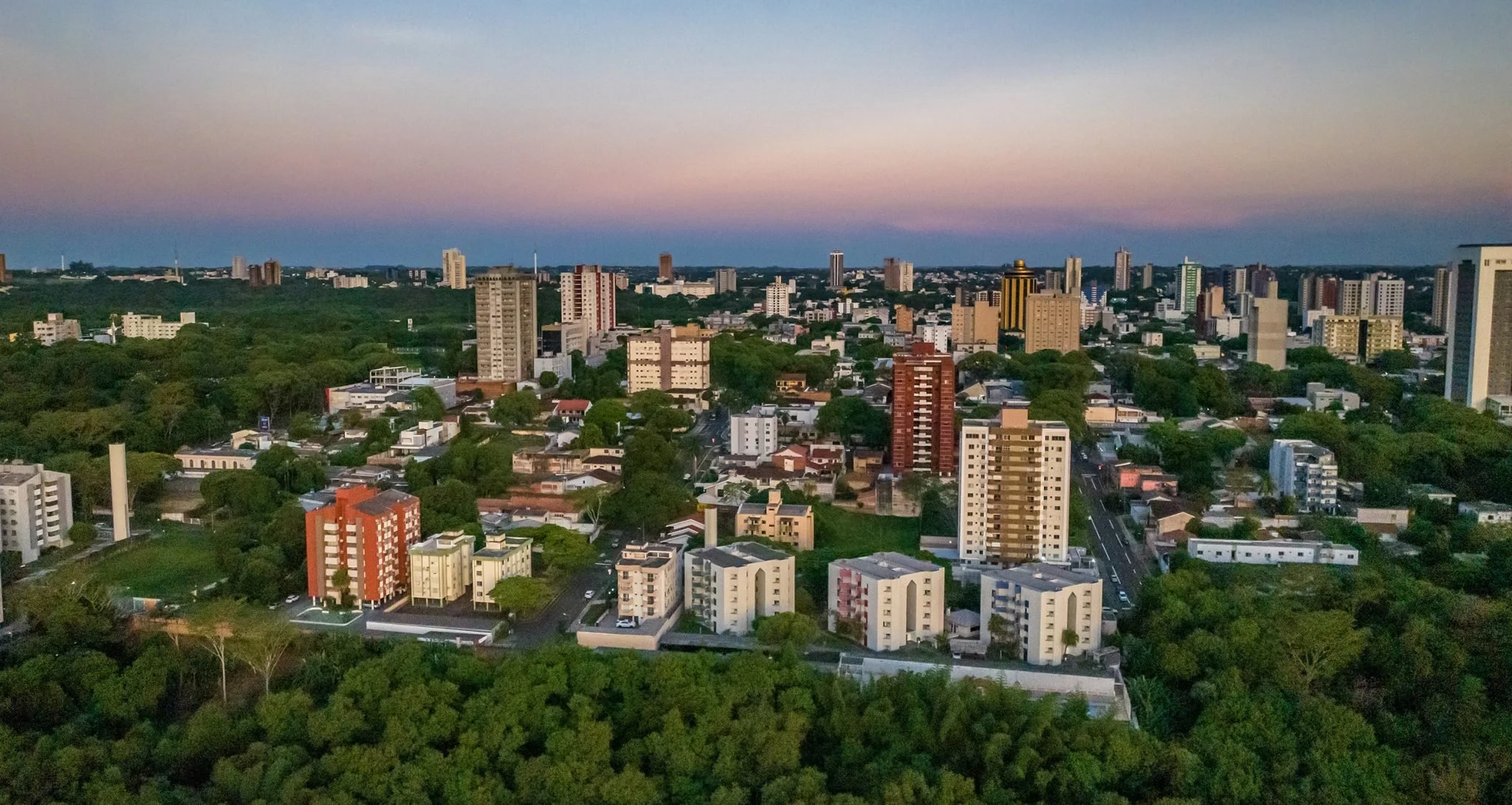 A cityscape at dawn with a mix of high-rise buildings and greenery, under a sky with pastel colors.