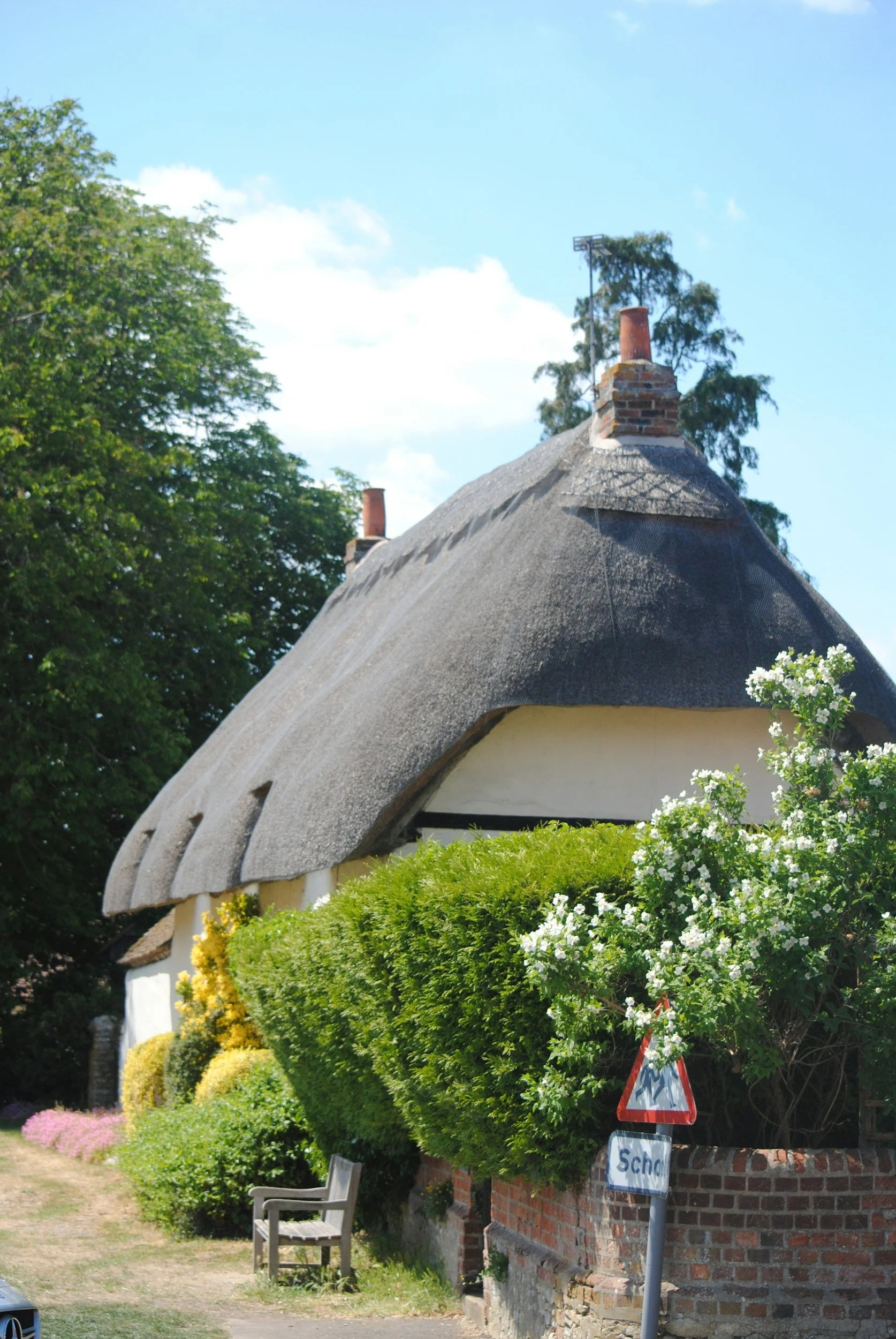 A traditional English cottage with a thatched roof, surrounded by green bushes and flowering plants, under a blue sky with clouds.