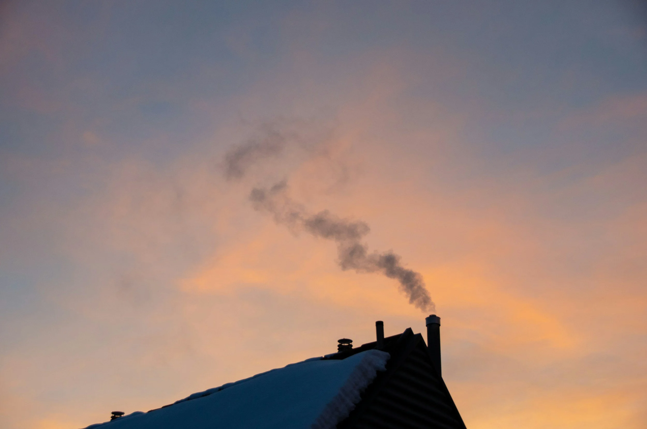 Smokestack emitting dark smoke against a colorful sunset sky, with a snow-covered rooftop in the foreground.