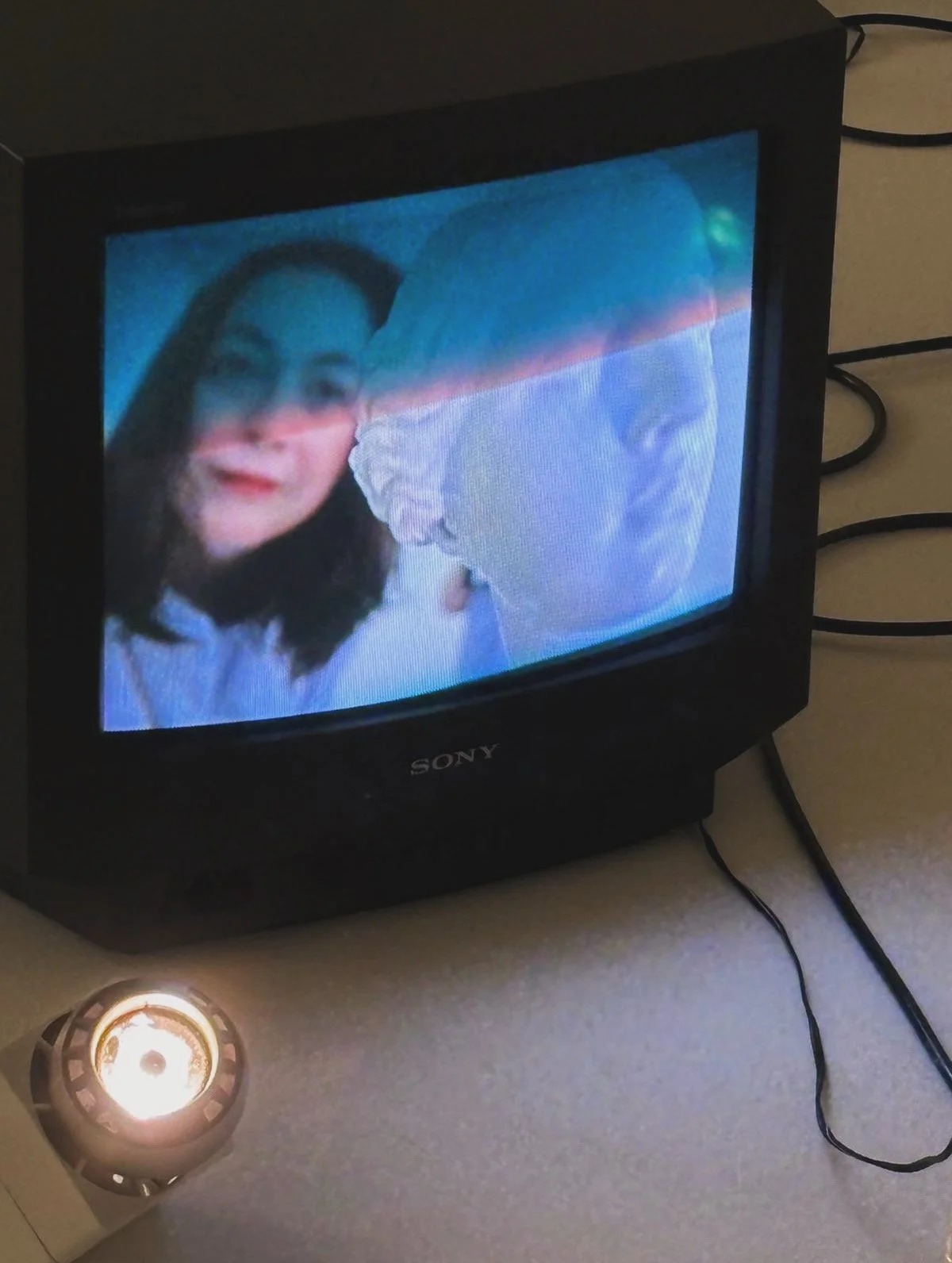 A small Sony television displaying an image of two people, one woman and one man, close-up. The woman has dark hair, and the man is wearing a white hat and glasses, both looking at the camera. There is a light source or candle placed on the table in front of the TV.