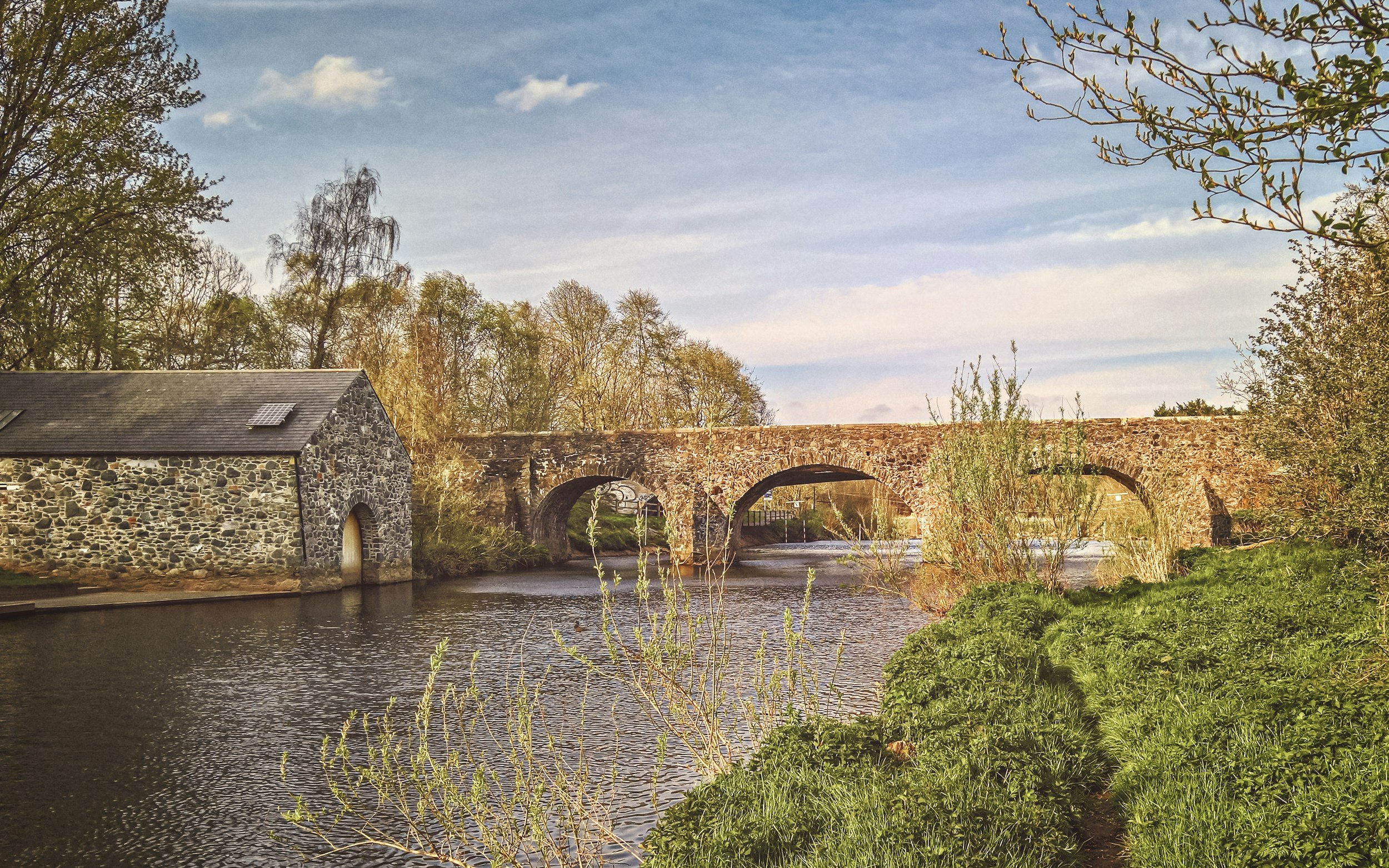 A stone building with a slate roof and a solar panel is situated by a river. An old stone bridge with arches spans across the river, surrounded by trees and greenery. The sky is partly cloudy with blue and white clouds.