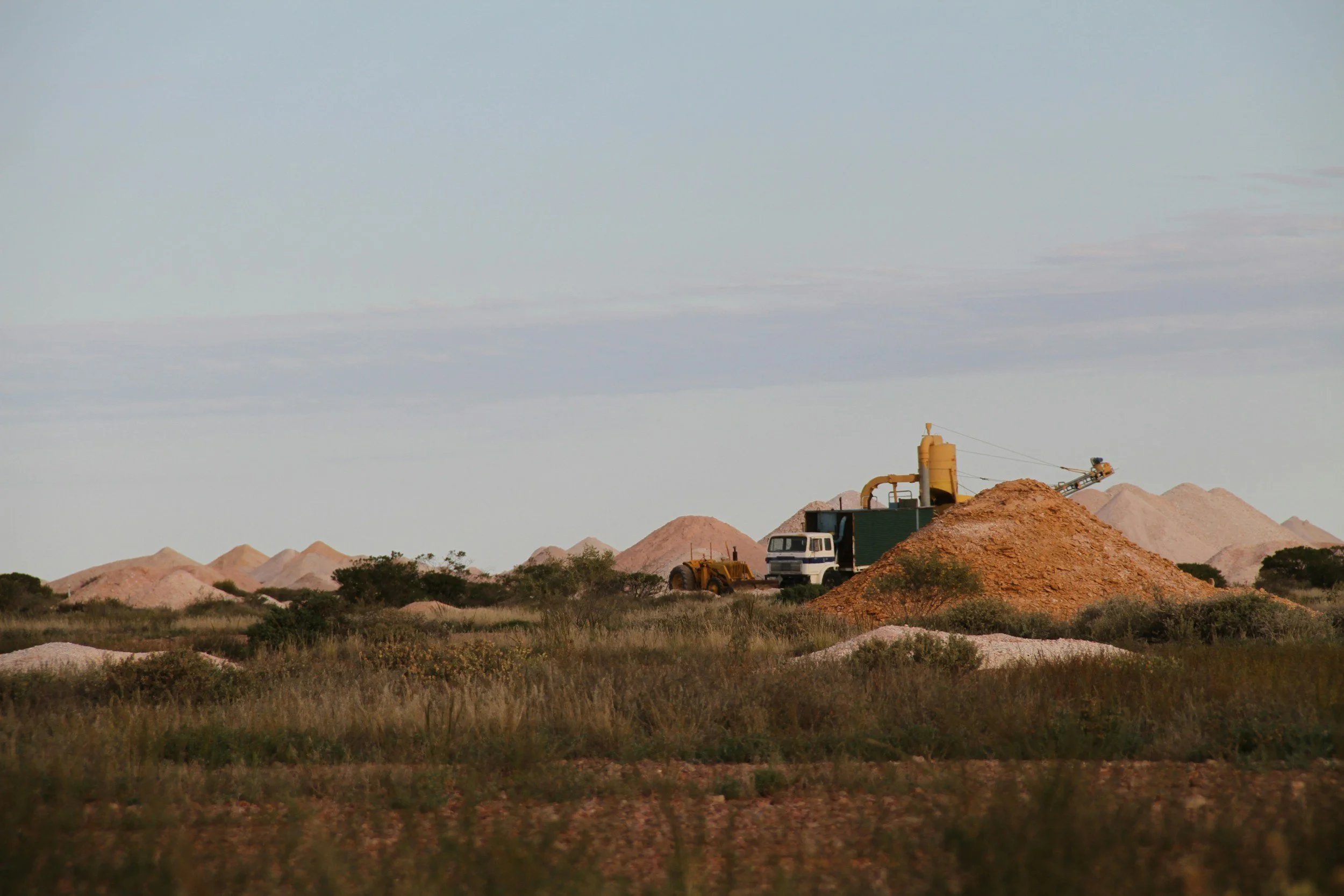 Construction site with yellow machinery, a white vehicle, and mounds of dirt in a semi-arid landscape with hills and sparse vegetation under a mostly cloudy sky.