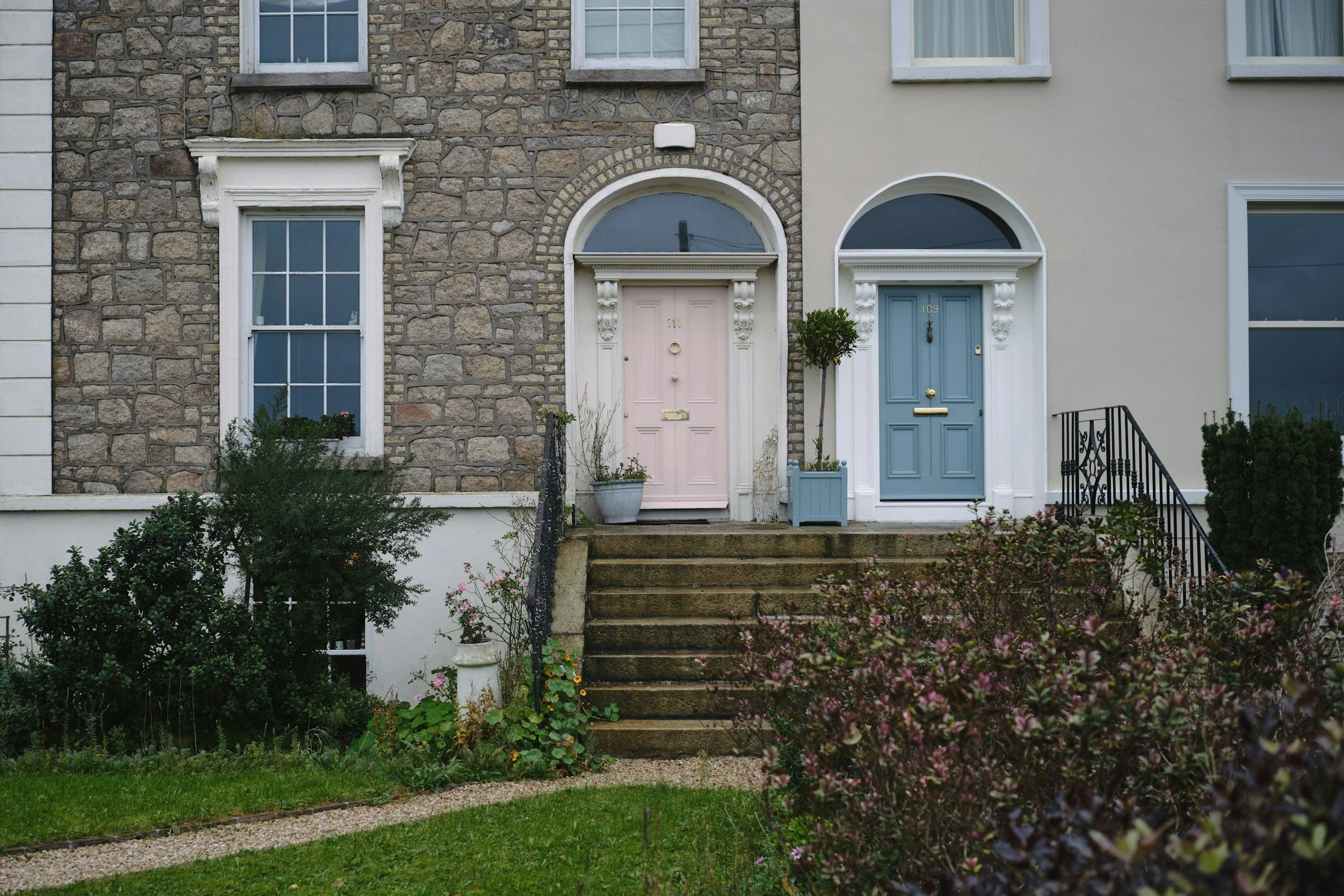 Front view of two adjacent houses with different exterior styles, one with stonework and one with smooth plaster, showing front doors, windows, stairs, plants, and greenery.