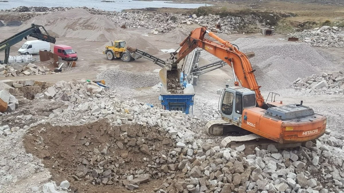 Construction site with a large orange Hitachi excavator loading rocks into a blue dumpster, surrounded by piles of rocks and construction vehicles.