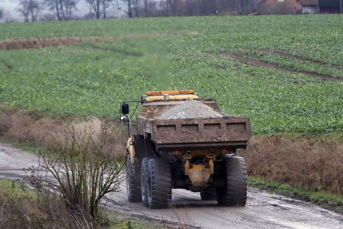 Large yellow dump truck driving on a dirt road in a rural area with green fields and sparse trees in the background.