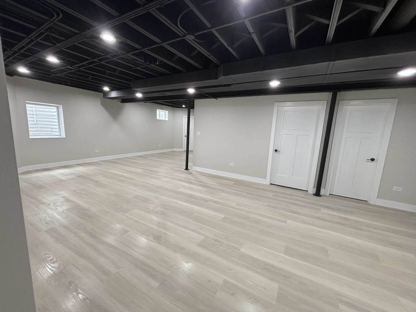 Empty basement room with light-colored wood flooring, white walls, two small windows, black ceiling with exposed beams and recessed lighting, white doors, and a black support column.