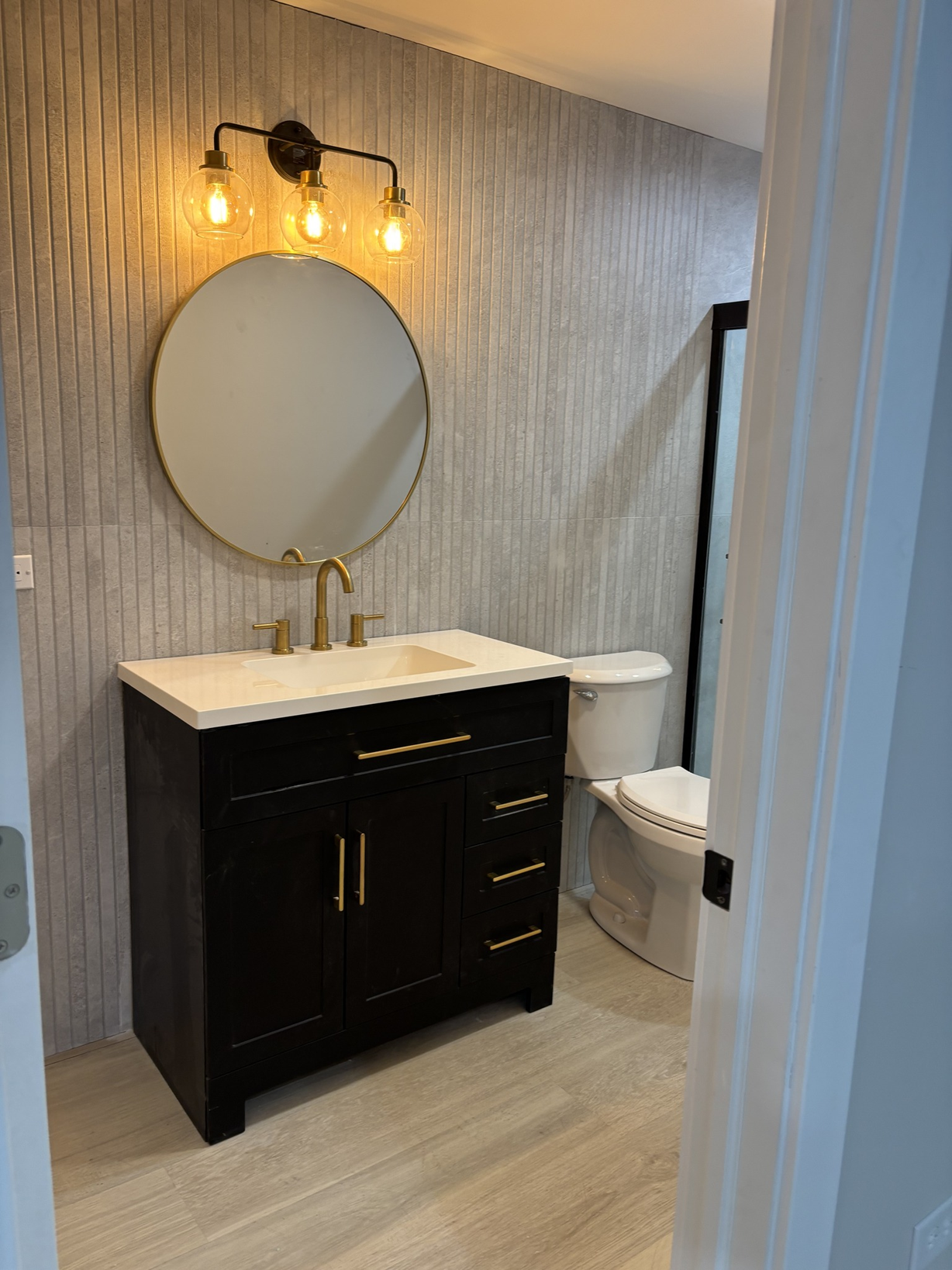 Modern bathroom with a round mirror, black vanity with gold handles, gold faucet, and a white toilet, all illuminated by warm vanity lights.