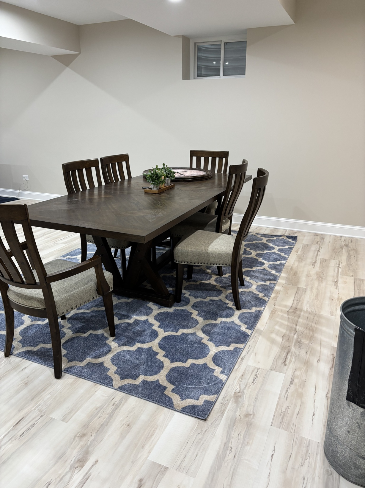 A dining room with a rectangular wooden table, six chairs with cushioned seats, a decorative plant, and a patterned blue and beige rug on light wood flooring. A small window on the beige wall and a trash bin in the corner are also visible.