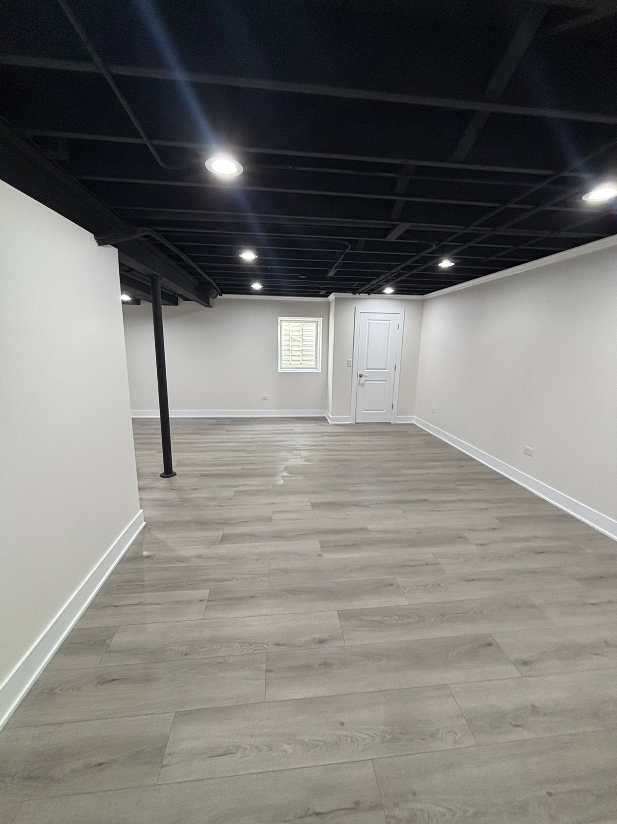 Empty basement room with light gray wood flooring, white walls, black ceiling with recessed lighting, a small window, and a white door.