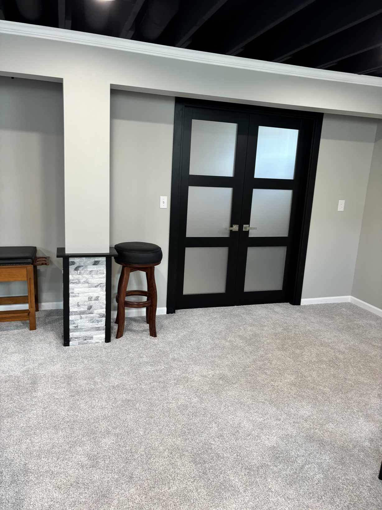 Interior room with light gray walls, beige carpet, black double-door with frosted glass panels, and a small bar area with a wooden stool and a short black table with a stone facade.