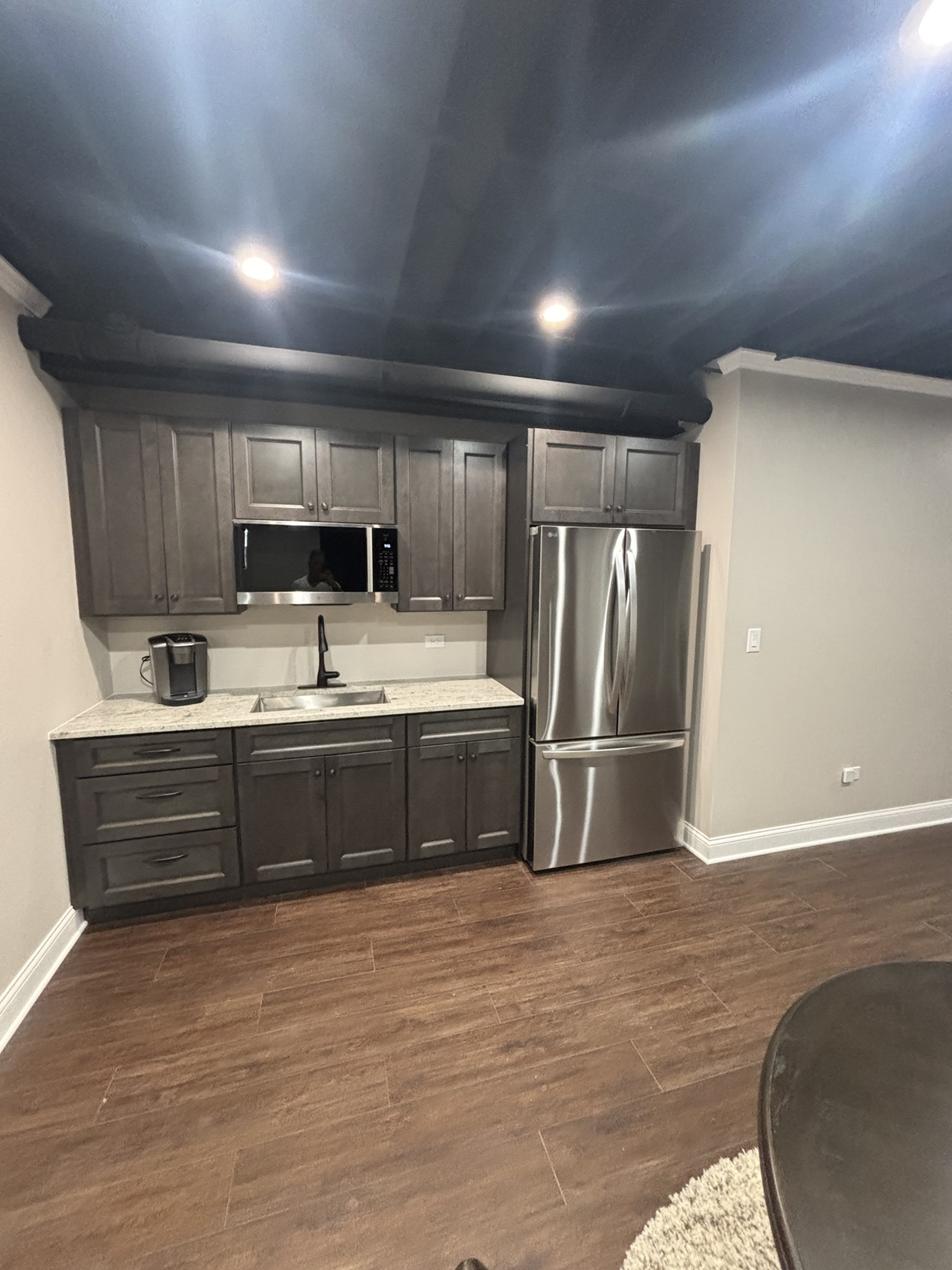 Modern kitchen with dark wood cabinets, stainless steel refrigerator, microwave, granite countertop, black faucet, and wood flooring.