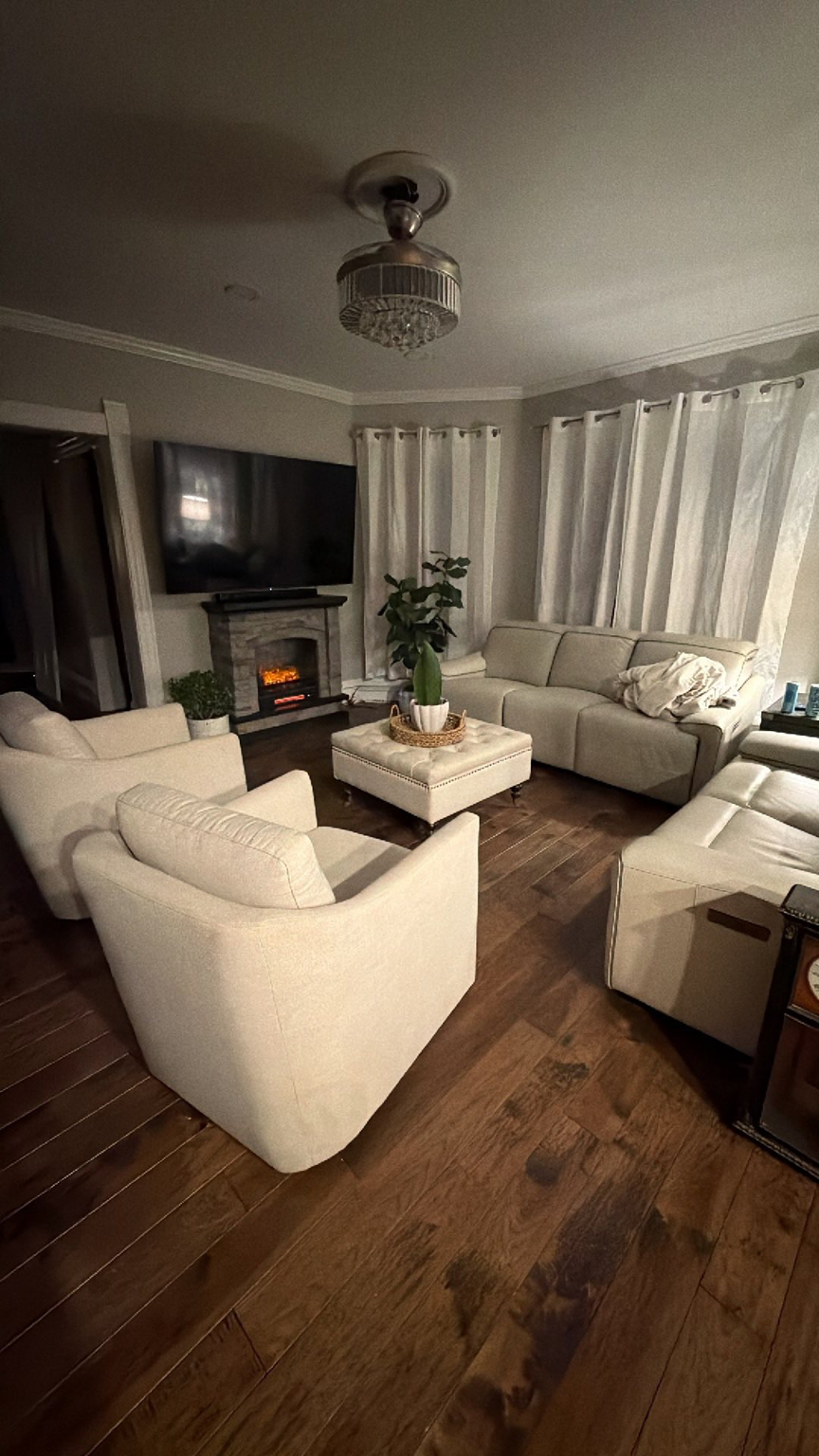 Living room with beige furniture, a fireplace, a large TV, and a plant, with hardwood floors and white curtains.
