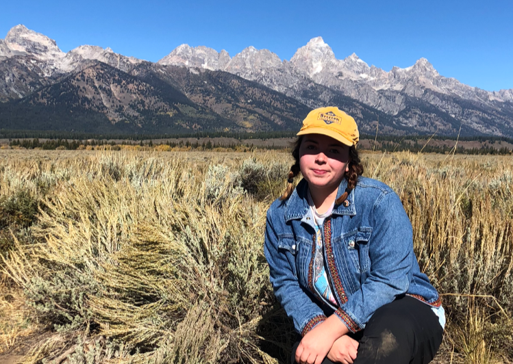 A young girl sitting in a grassy field with mountains in the background, wearing a yellow cap and denim jacket.