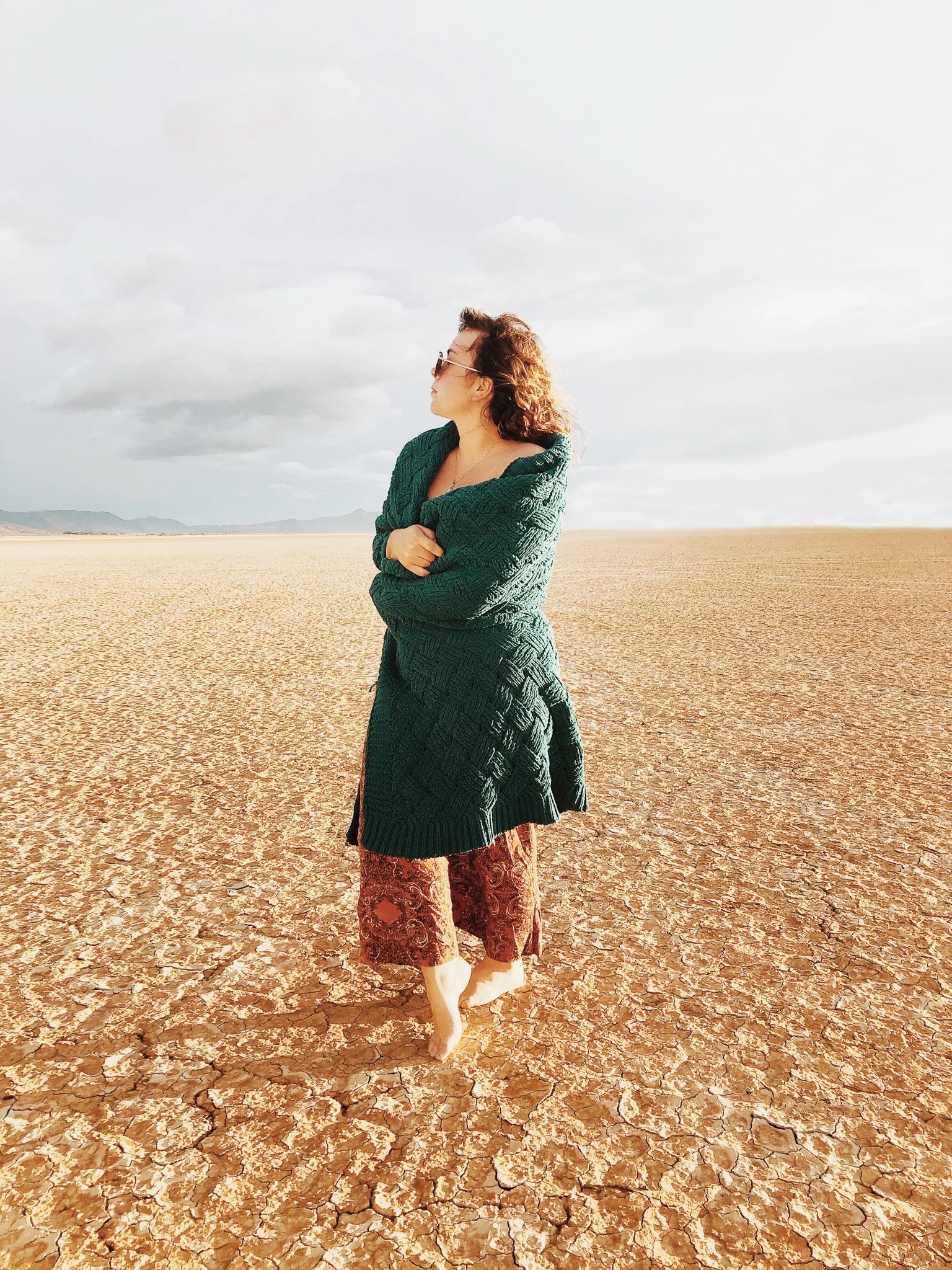 Woman with curly hair and sunglasses wearing a green sweater, patterned skirt, and patterned pants standing in a dry, cracked desert landscape under cloudy sky.