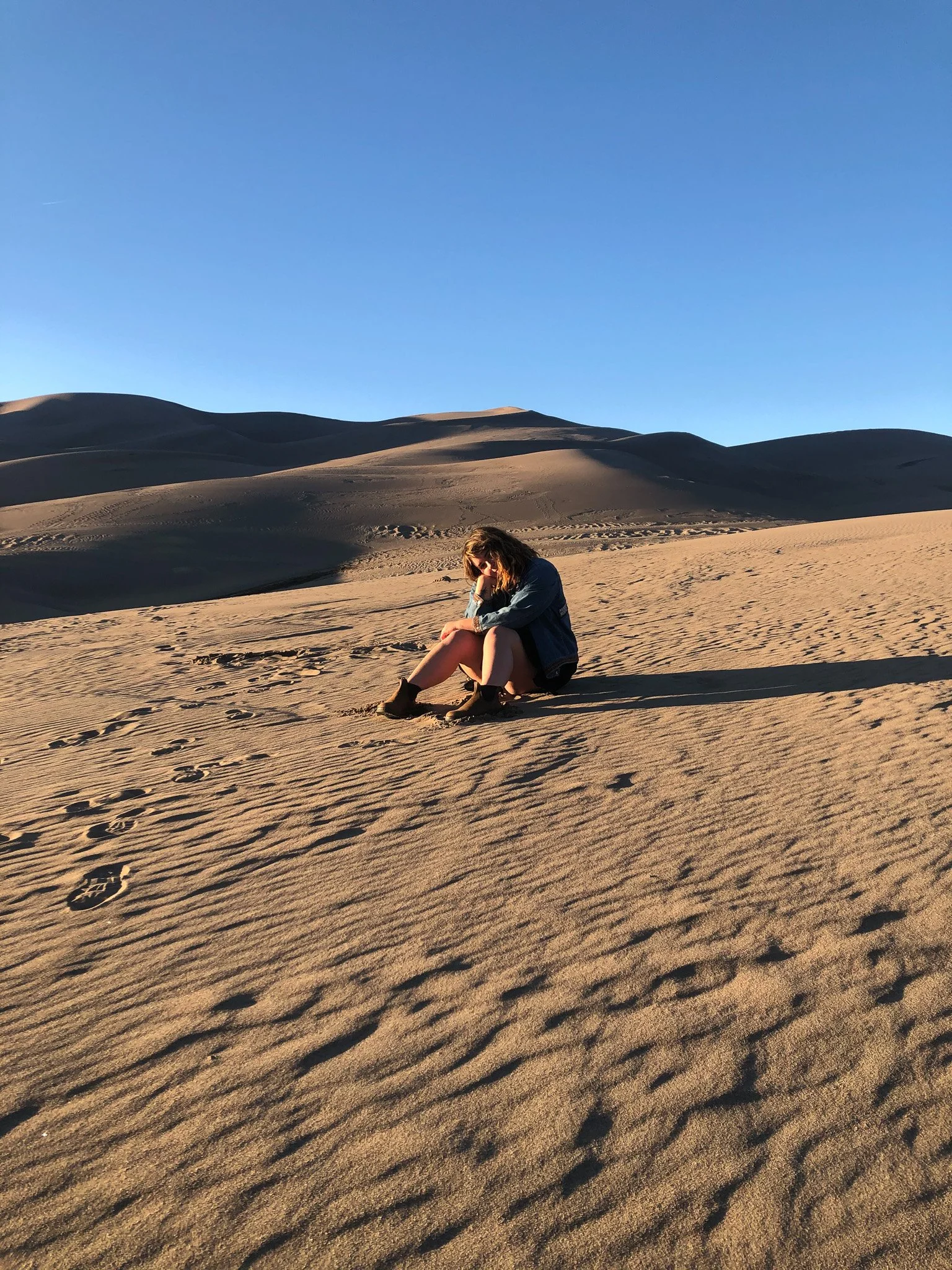 A woman sitting on the sand in a desert with sand dunes in the background and footprints in the foreground.