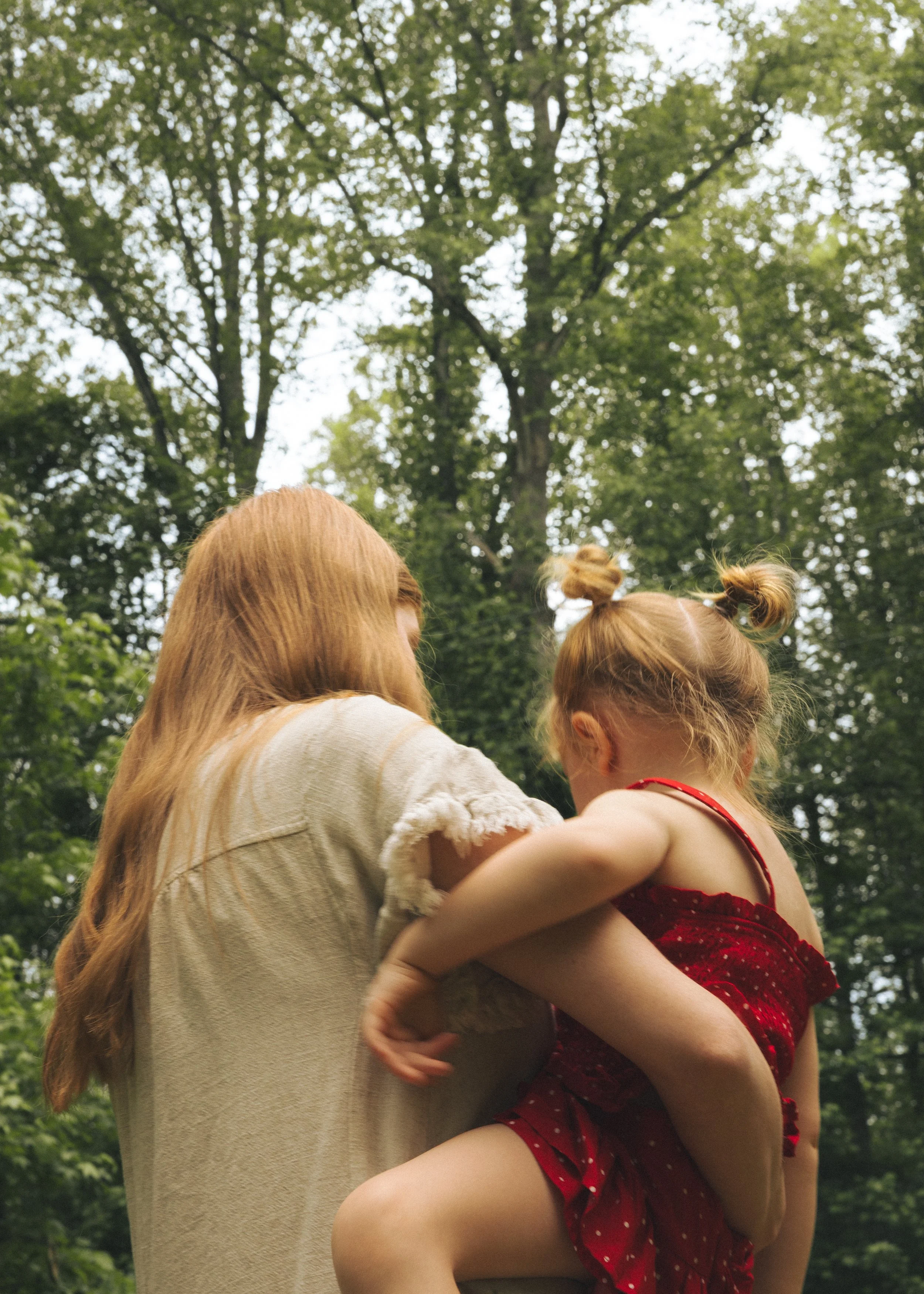 A woman holding a young girl in a red dress with white polka dots outdoors in a wooded area, with large green trees and bright sky in the background.