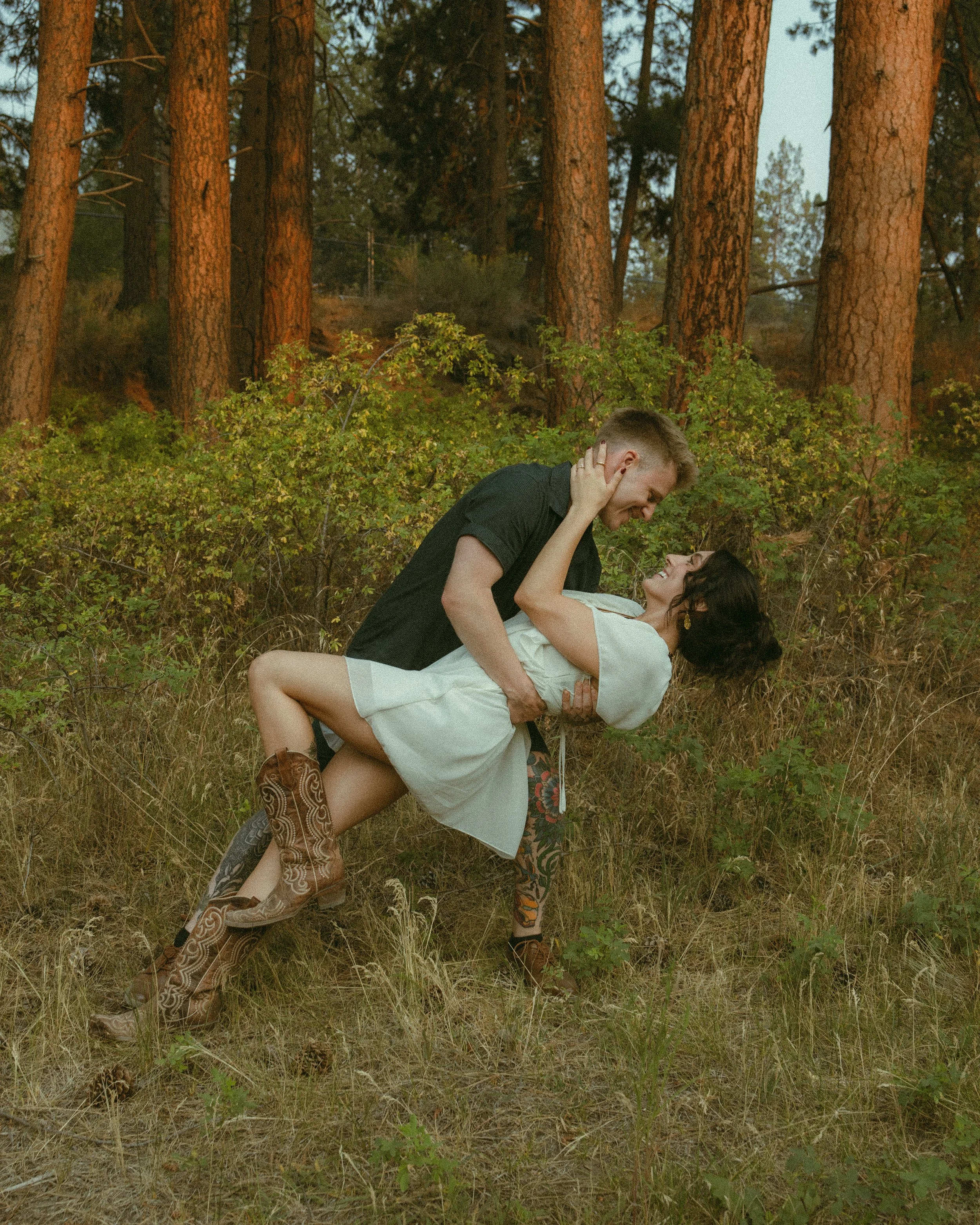 A man and woman lying on the ground in a forest, smiling and looking at each other, with the man holding the woman and their faces close together.