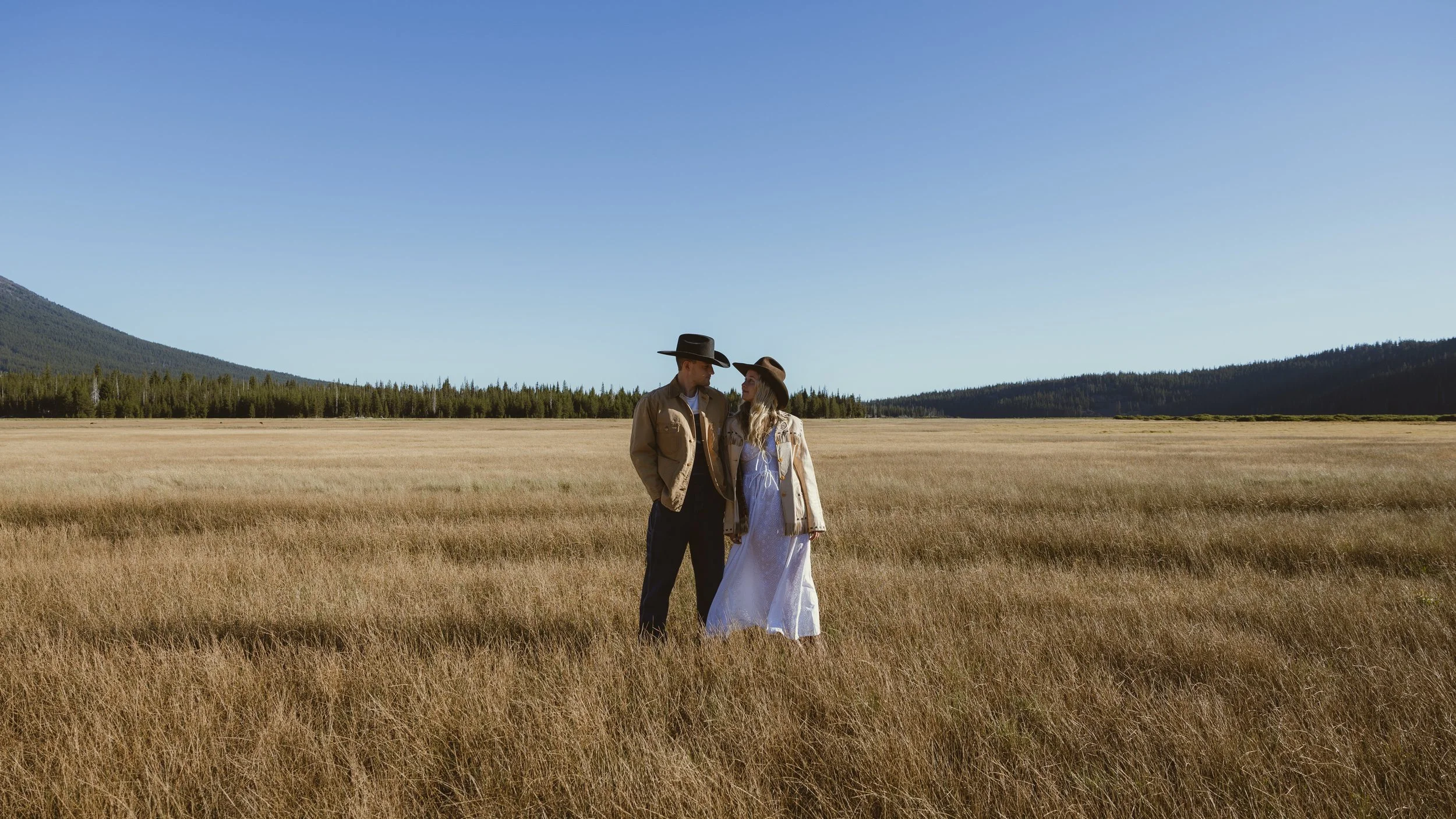 A couple dressed in country-style clothing standing in a golden grassy field with mountains and a clear blue sky in the background.